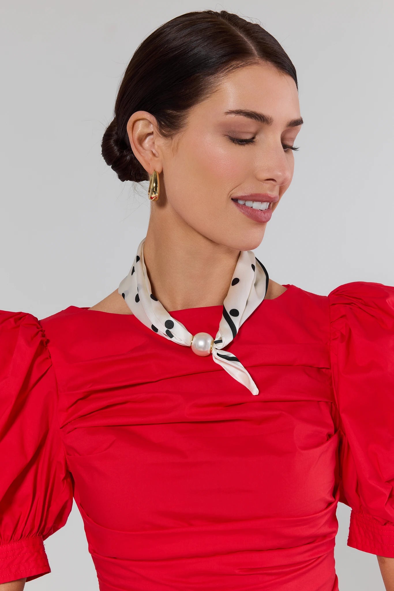 Woman wearing a red dress with a polka dot scarf and pearl necklace against a gray background