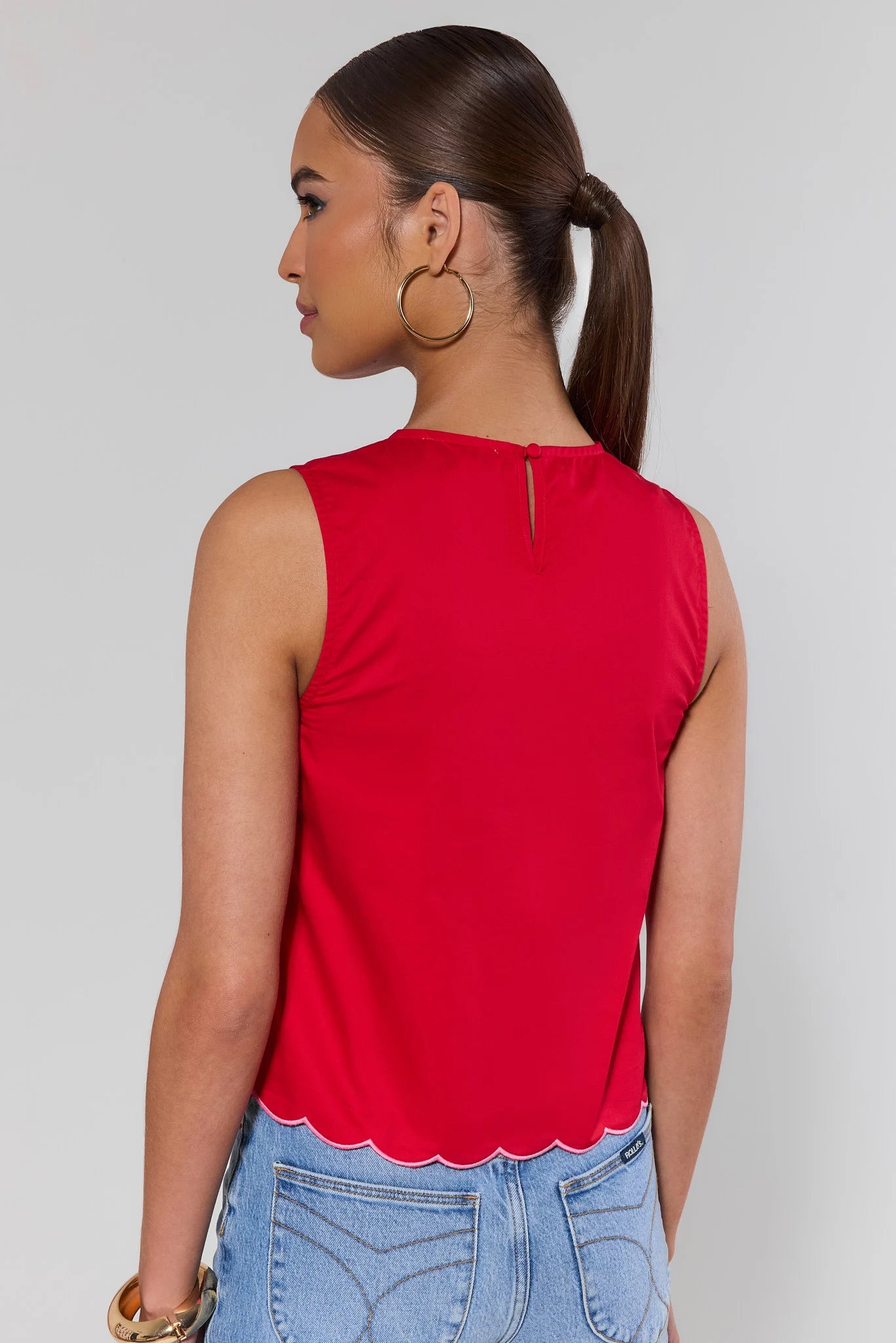 Woman standing wearing a red sleeveless top and blue jeans against a plain background