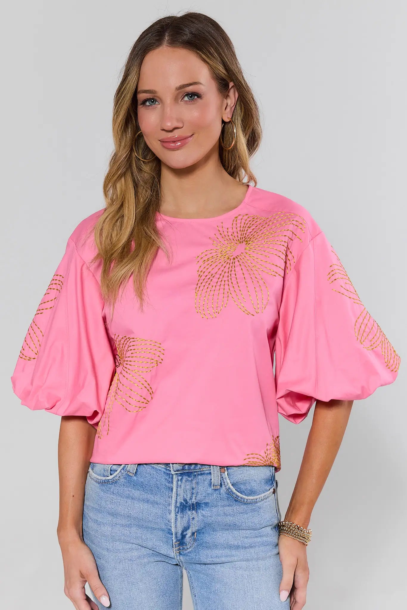 Woman wearing a pink blouse with floral embellishments and blue jeans on a plain background