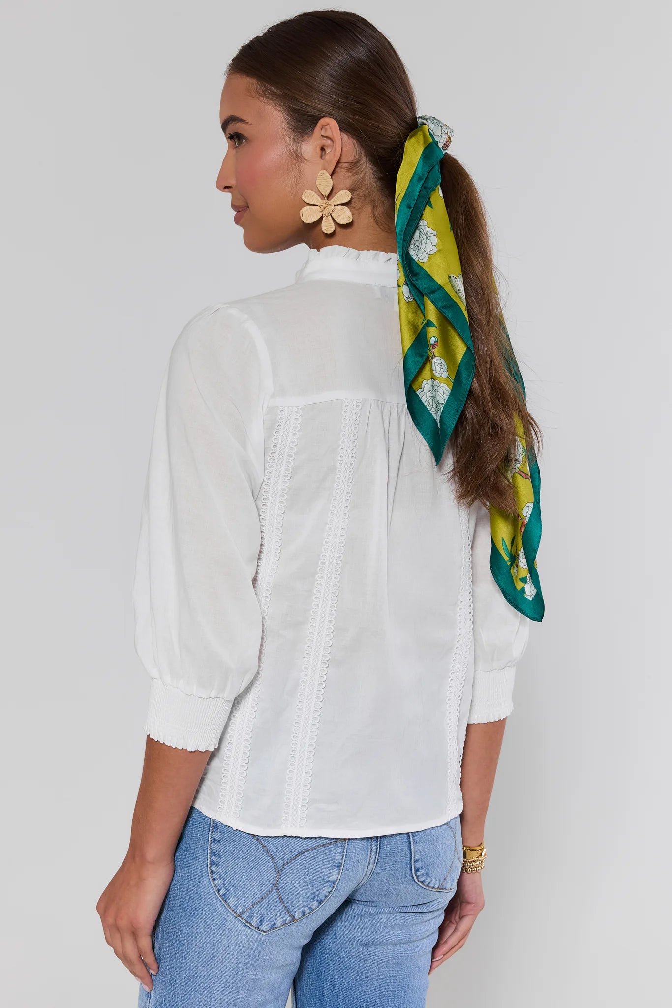 Woman wearing a white blouse and blue jeans with a green and yellow scarf on her head against a plain background