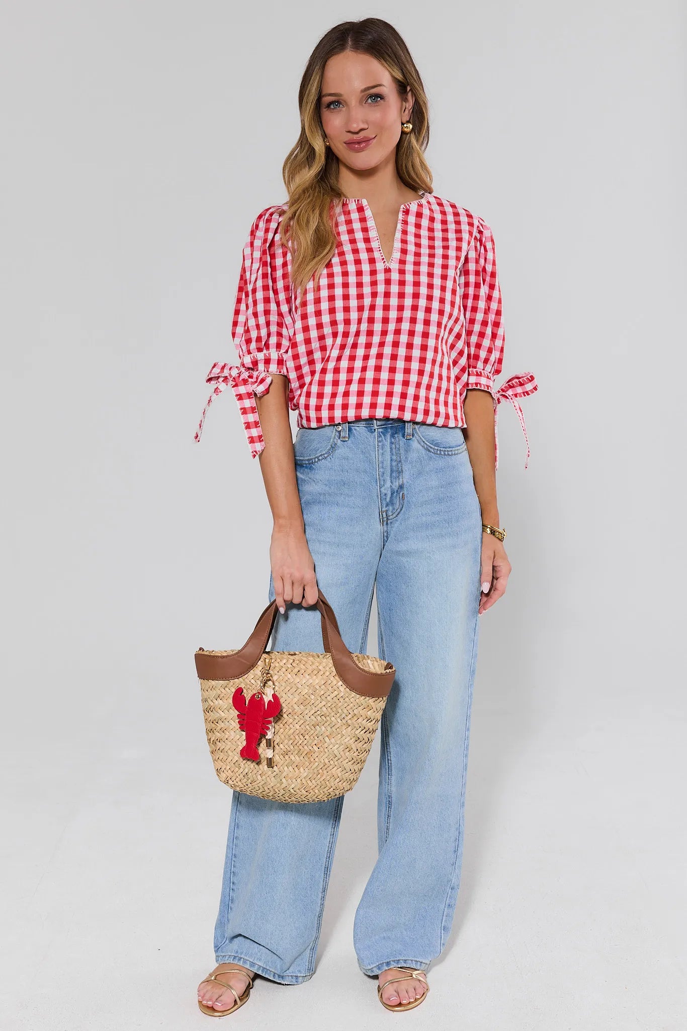 Woman wearing a red and white checkered blouse and blue jeans, holding a straw bag with a lobster design.