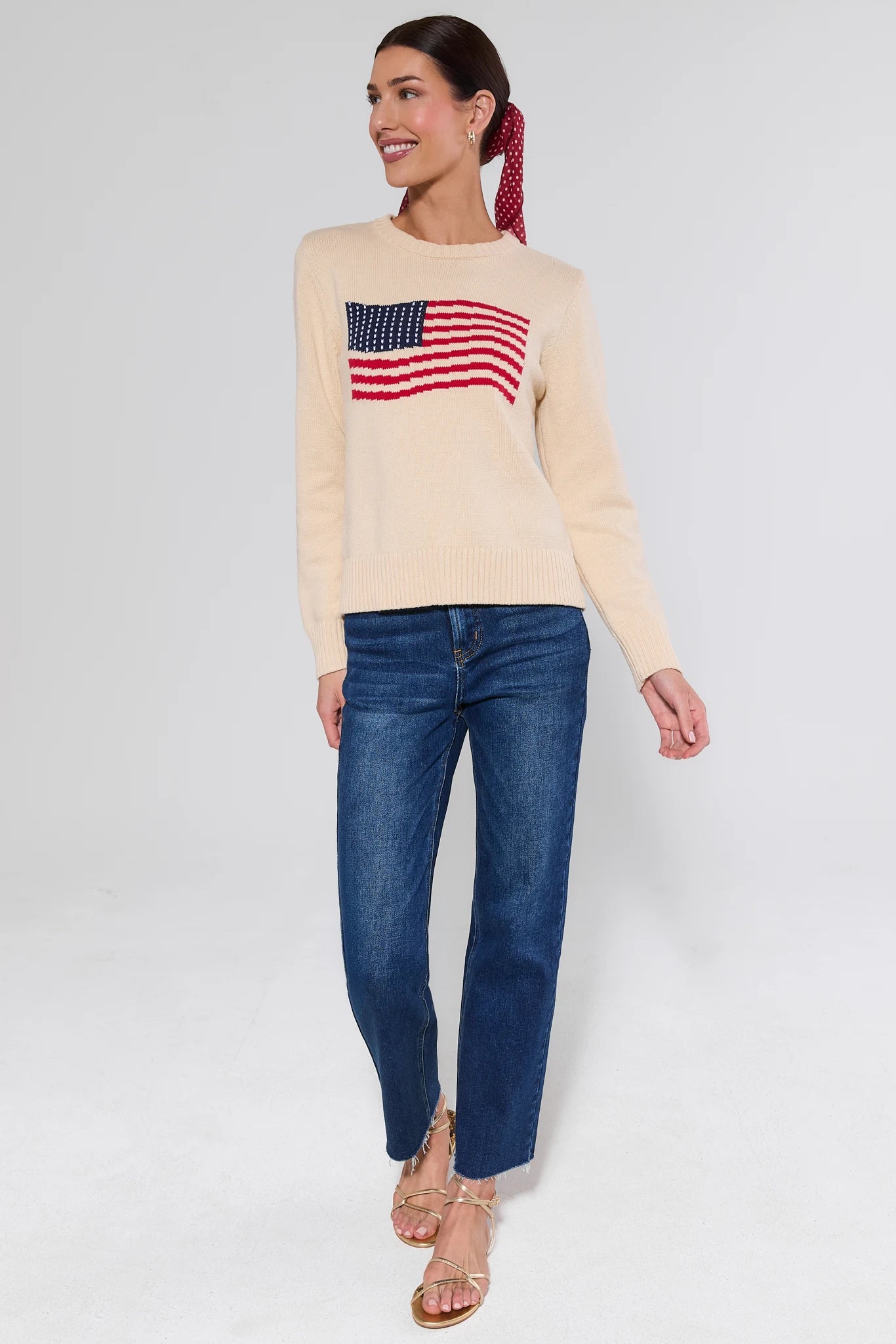 Woman wearing a beige sweater with an American flag design and blue jeans on a white background