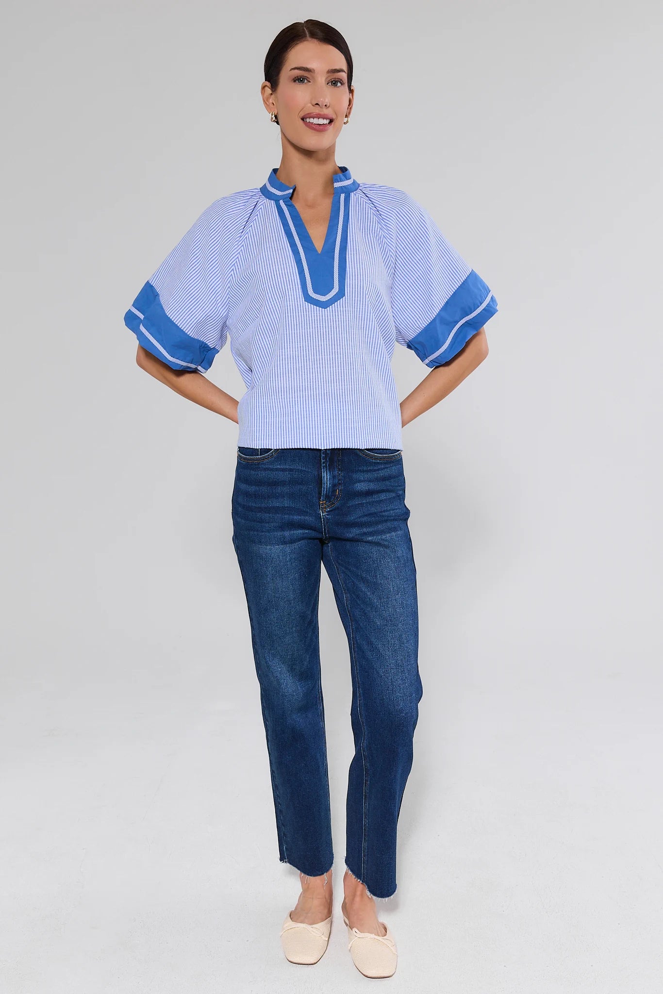 Woman wearing a light blue top with blue trim and dark blue jeans on a gray background