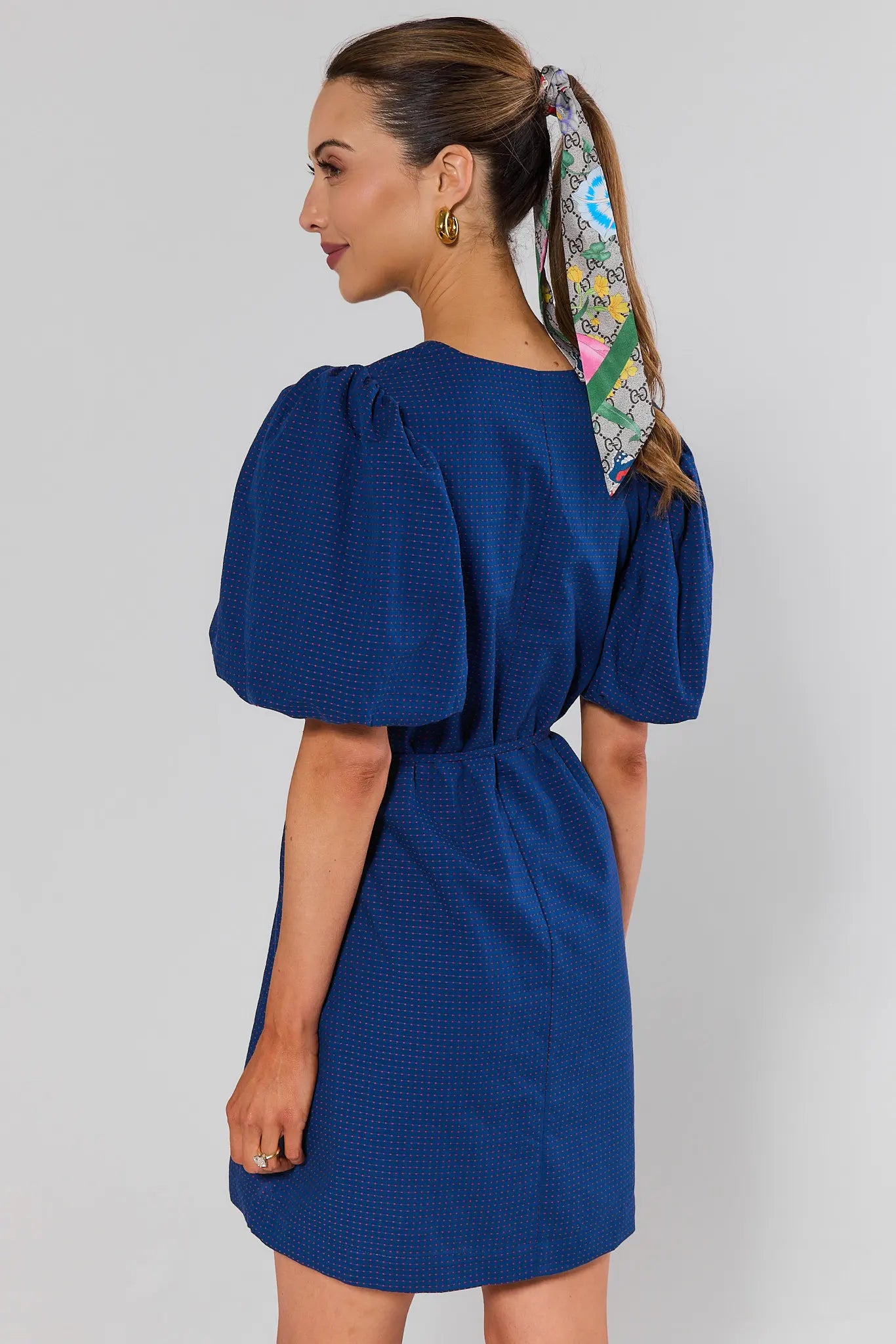 Woman standing wearing a blue dress with puff sleeves and a colorful scarf in her hair on a gray background