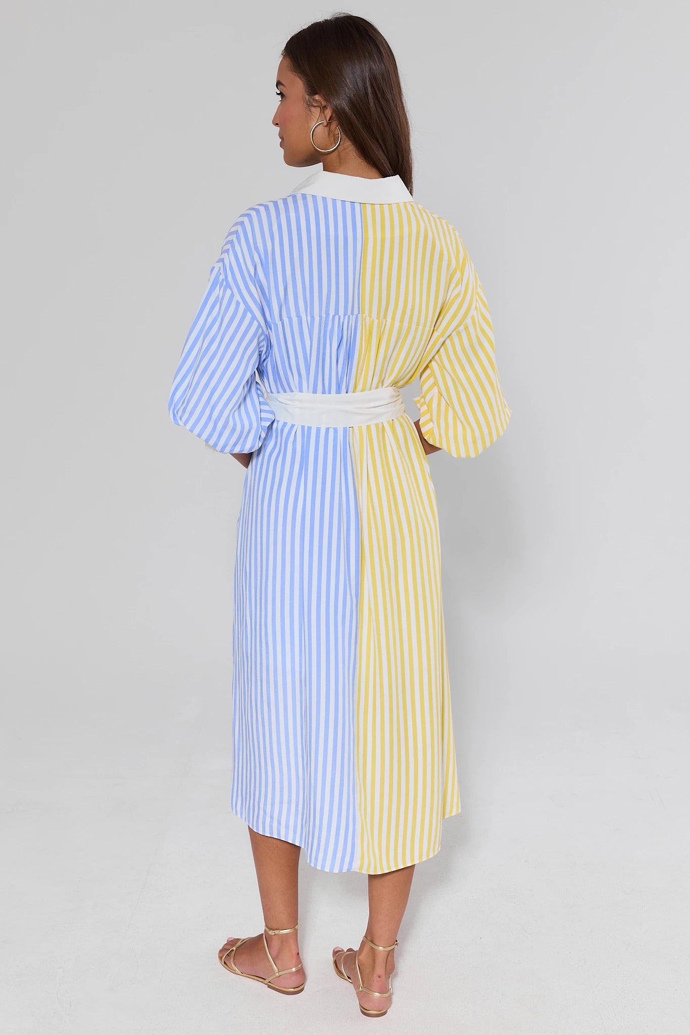 Woman standing wearing a colorful striped dress with a white belt on a plain background