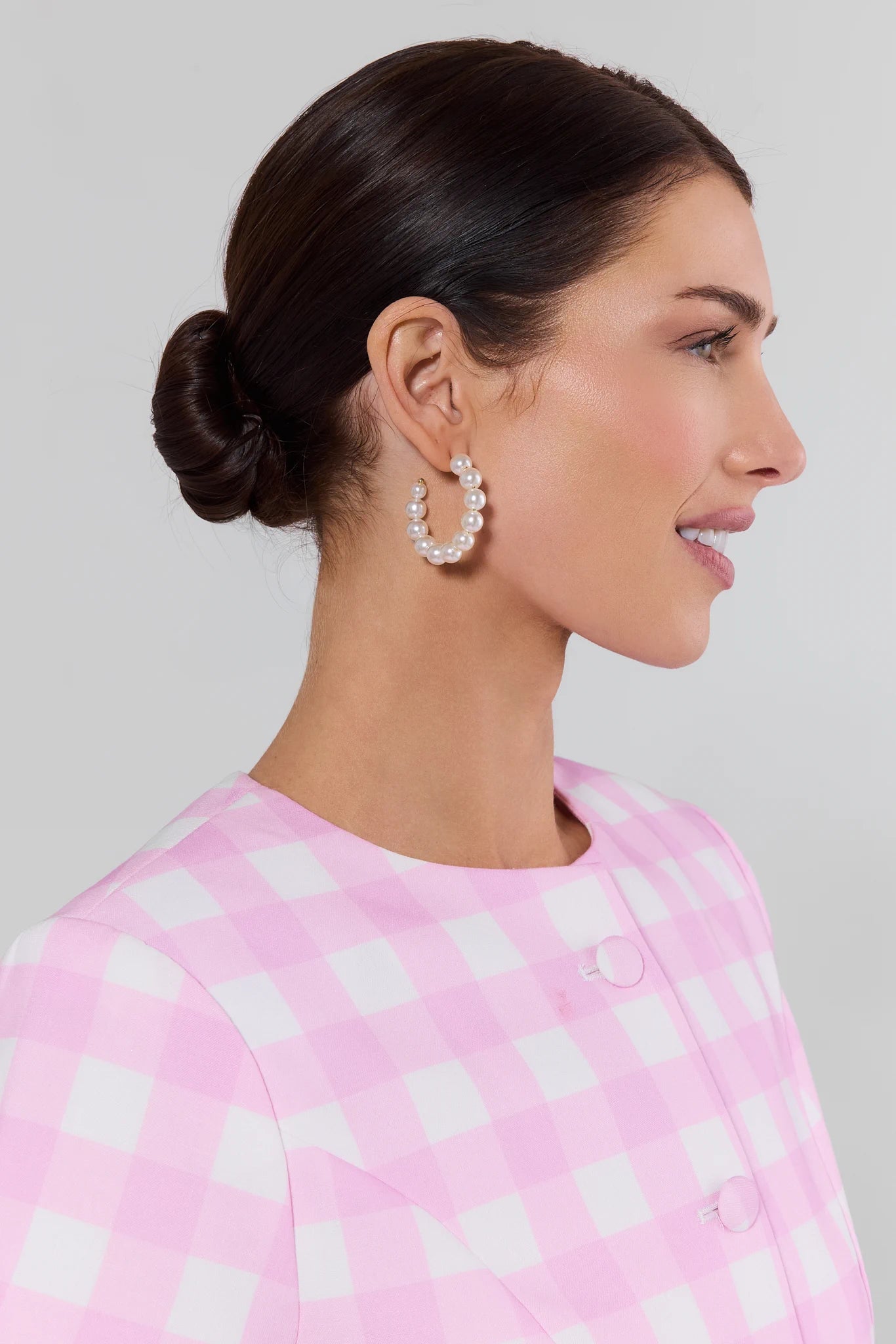 Woman wearing pearl hoop earrings with a pink and white checkered top against a light gray background