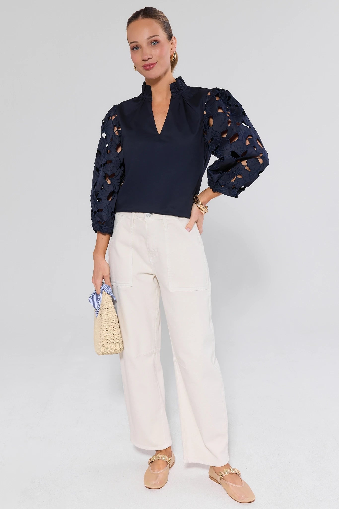 Woman wearing Navy Eyelet Sleeve Top by THML, featuring ruffled collar and voluminous sleeves, paired with high-waisted white trousers and a straw handbag.