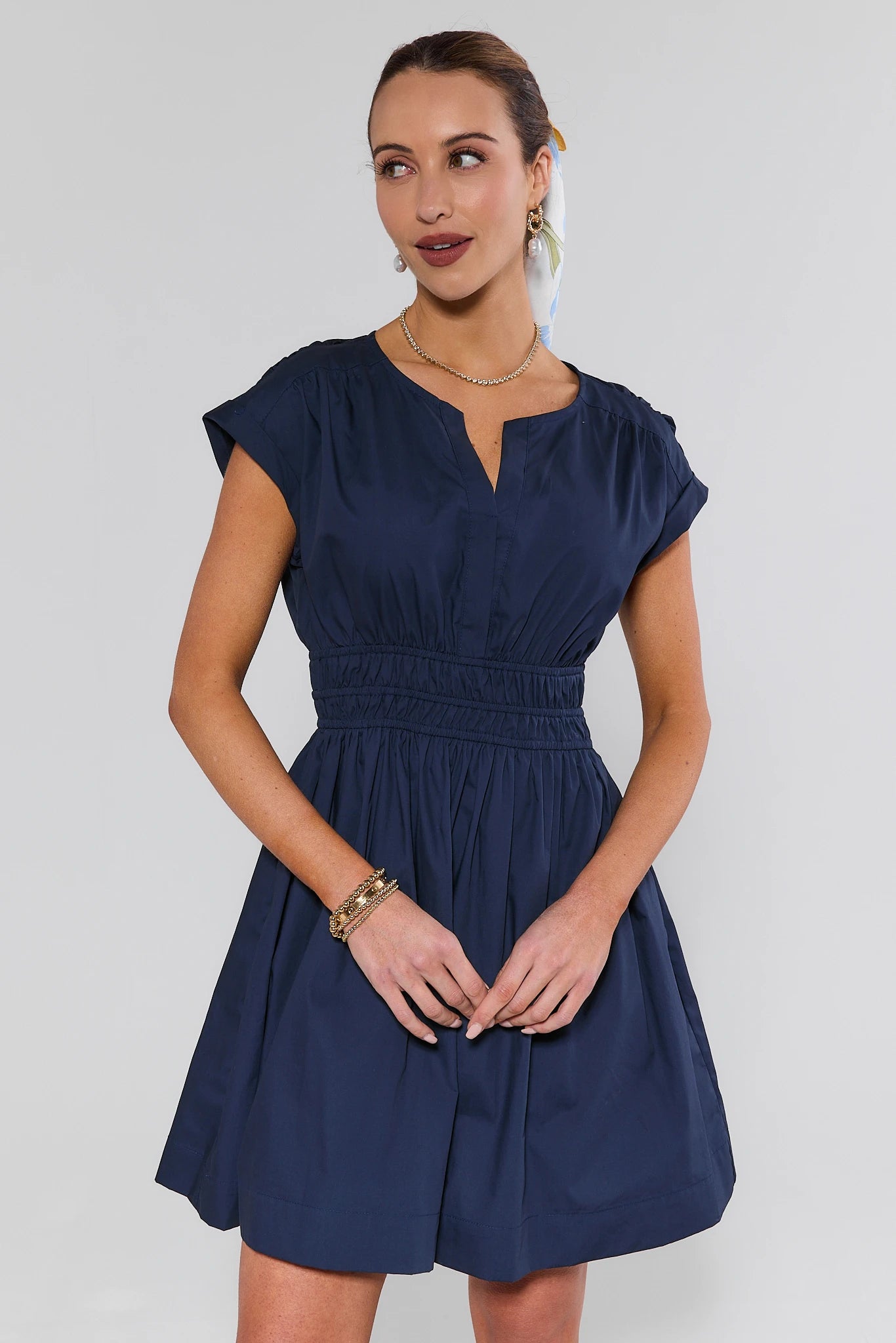 Model wearing Navy Smocked Waist Short Dress with a split-neckline, cap sleeves, and smocked waistband, accessorized with gold jewelry and a printed scarf.