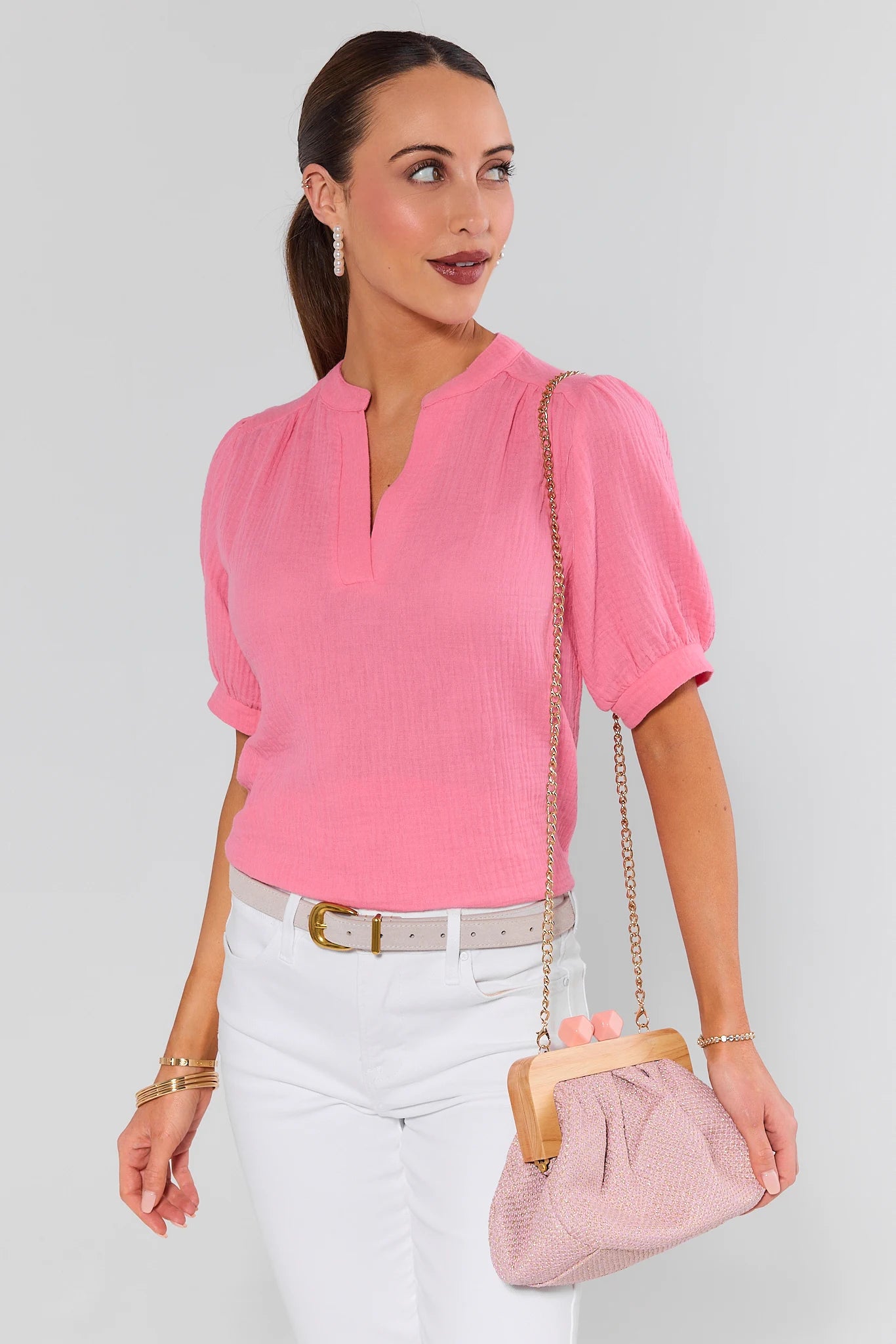 Woman wearing a pink blouse and white pants, holding a matching pink handbag against a plain background