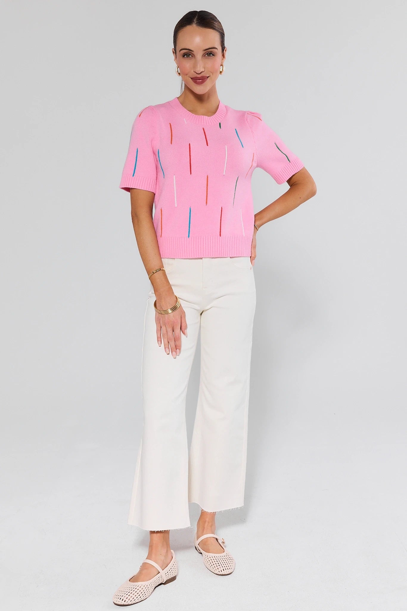 Woman wearing a pink top with abstract designs and white pants on a light gray background