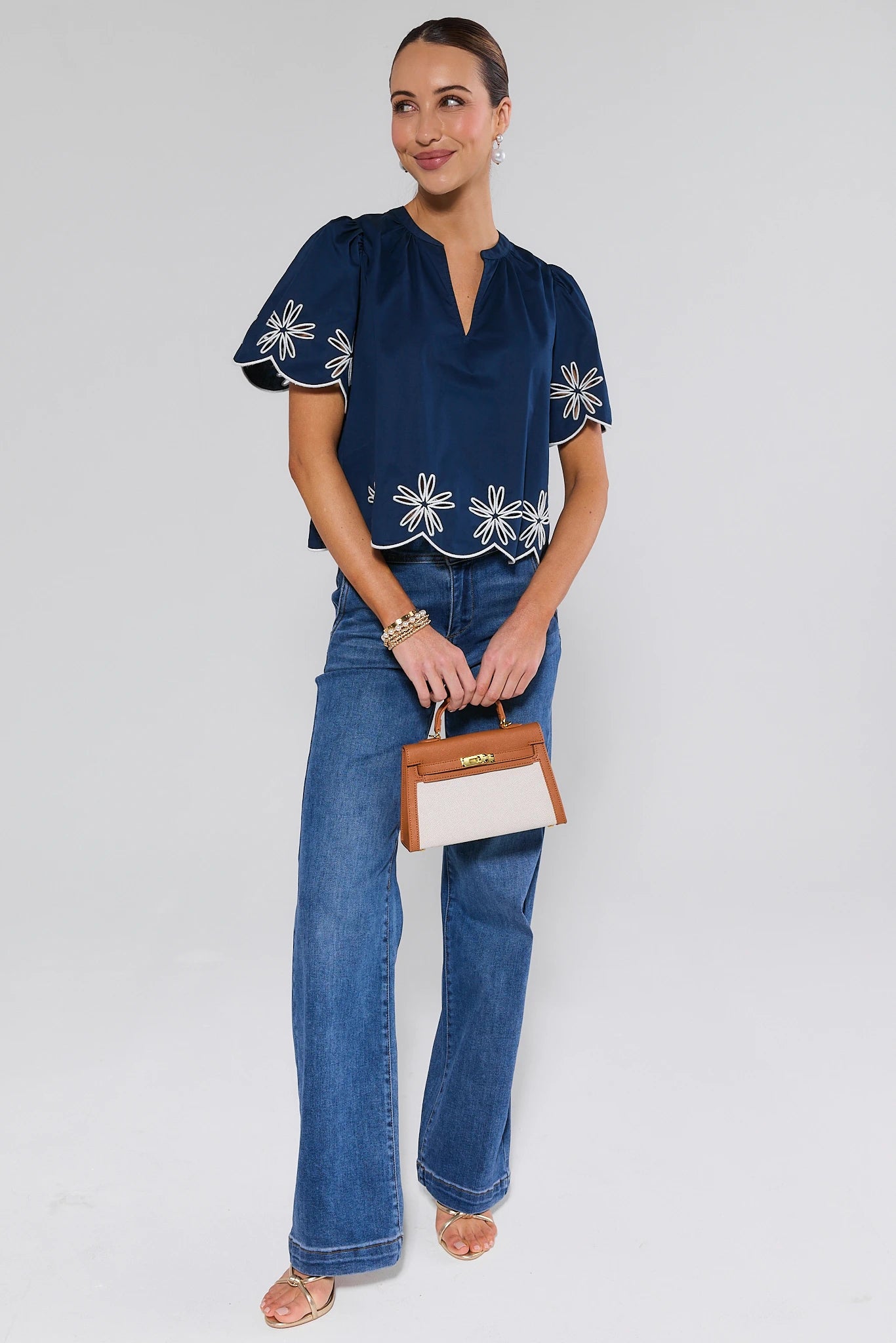 Woman wearing a navy blouse with white floral embroidery and blue jeans, holding a small handbag.