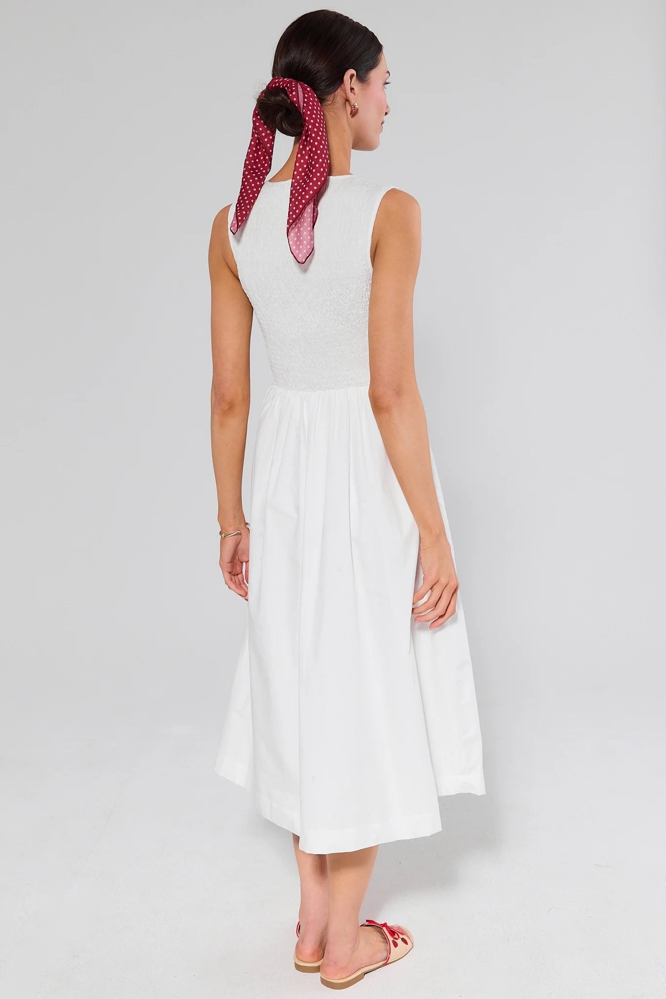 Woman standing wearing a white sleeveless dress with a red patterned headscarf on a plain background