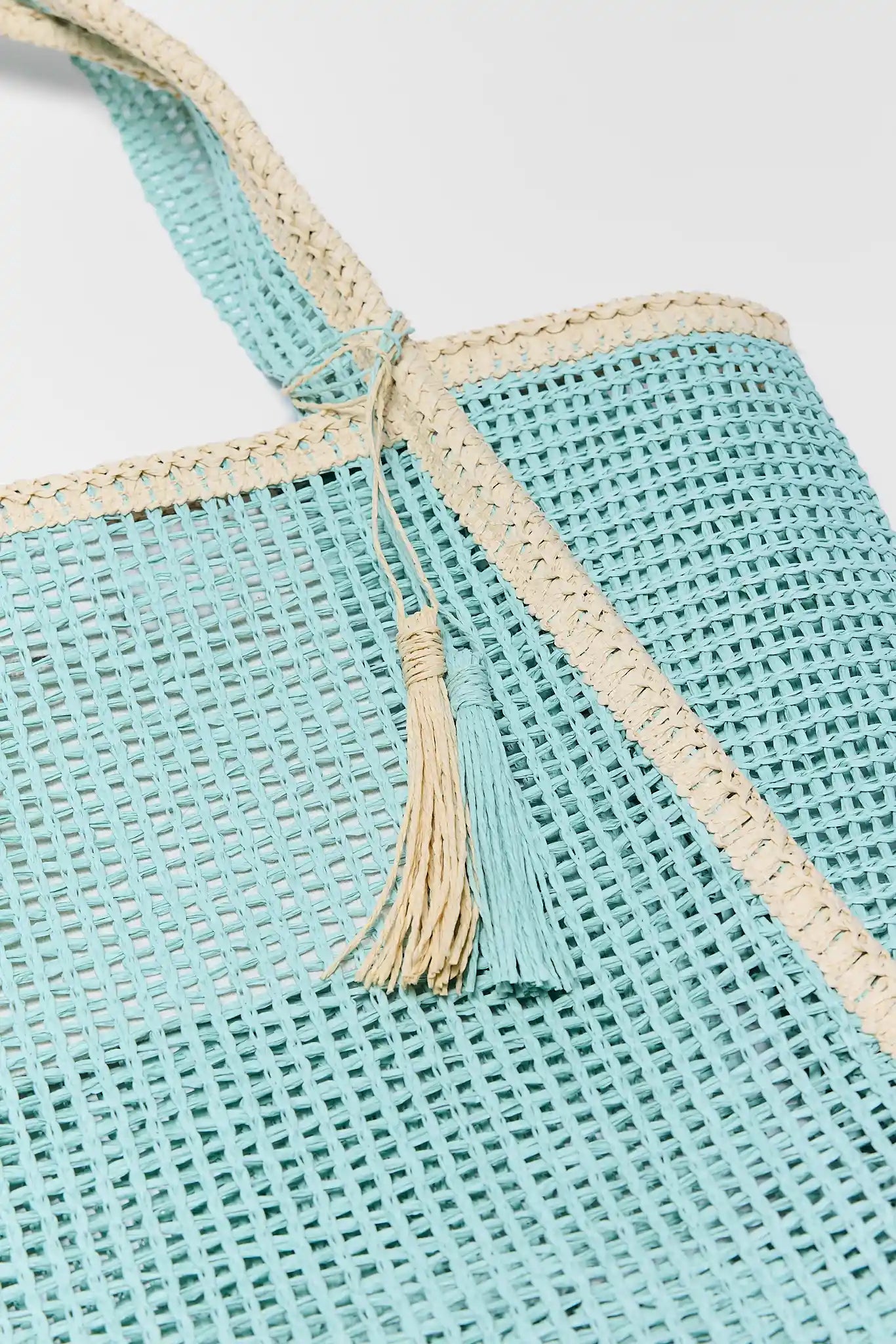 Close-up of a turquoise woven bag with beige straps and tassels on a light gray background