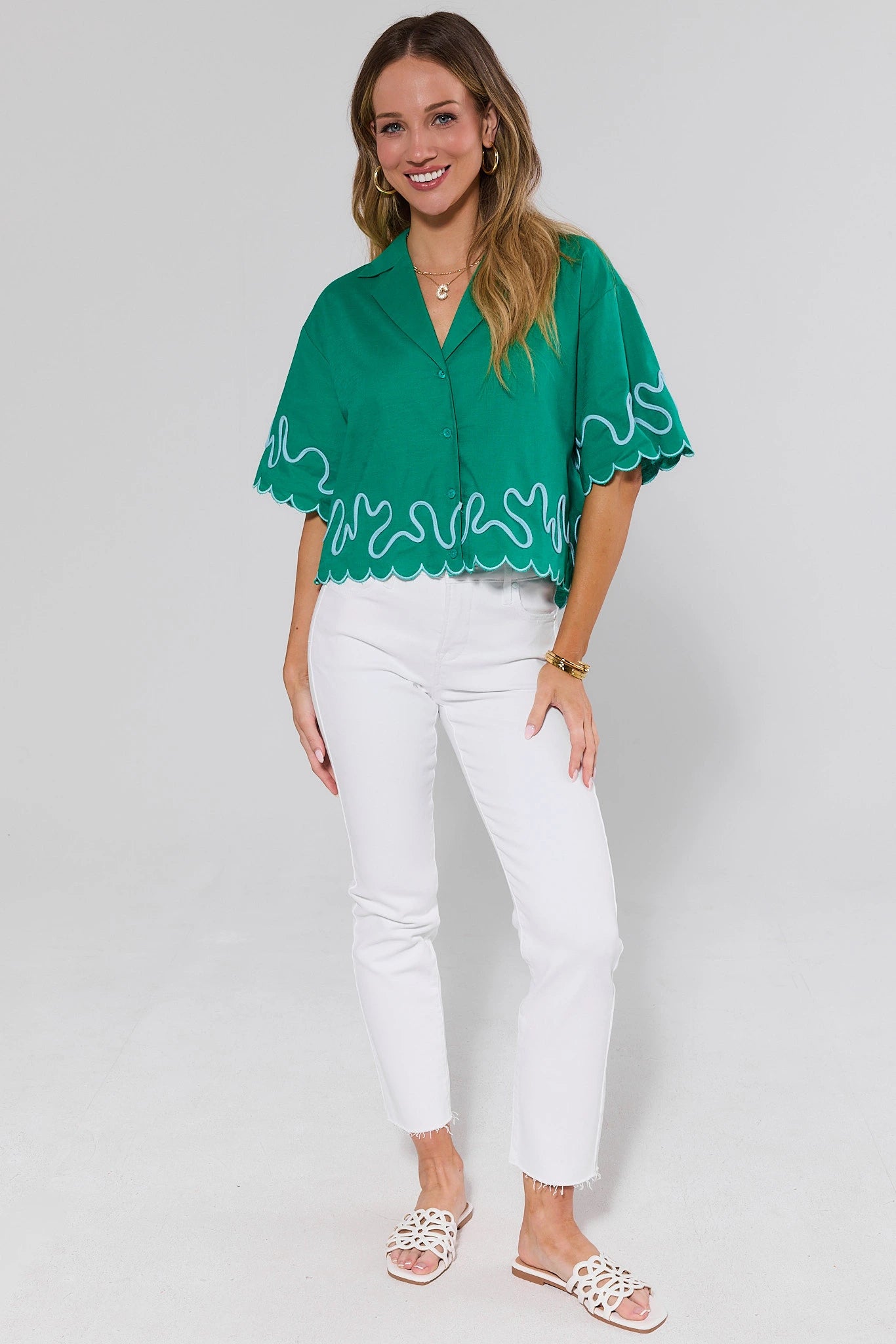 Woman wearing a green blouse with white trim and white pants on a light gray background