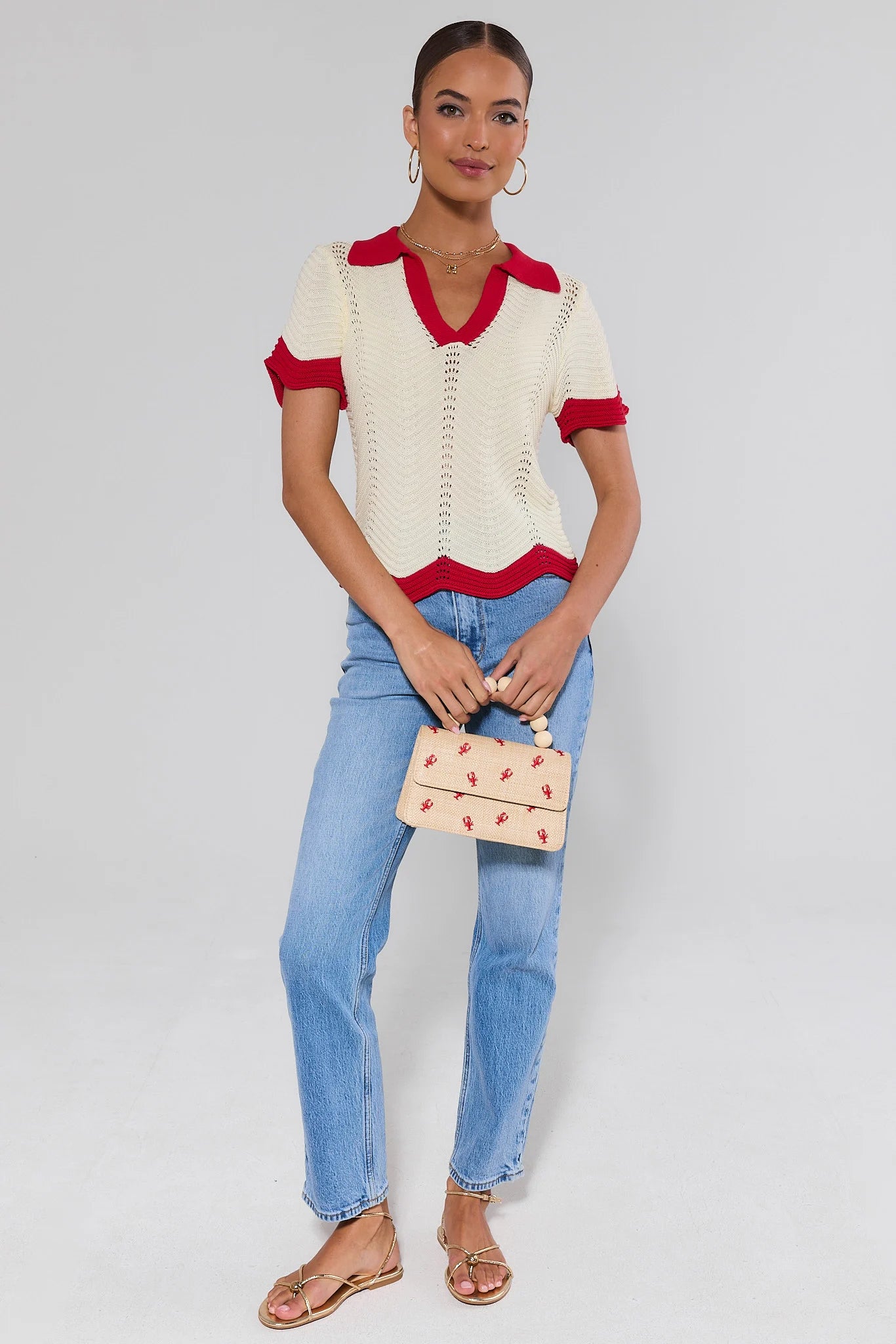 Woman wearing a ivory knit and red sweater with blue jeans, holding a small bag.