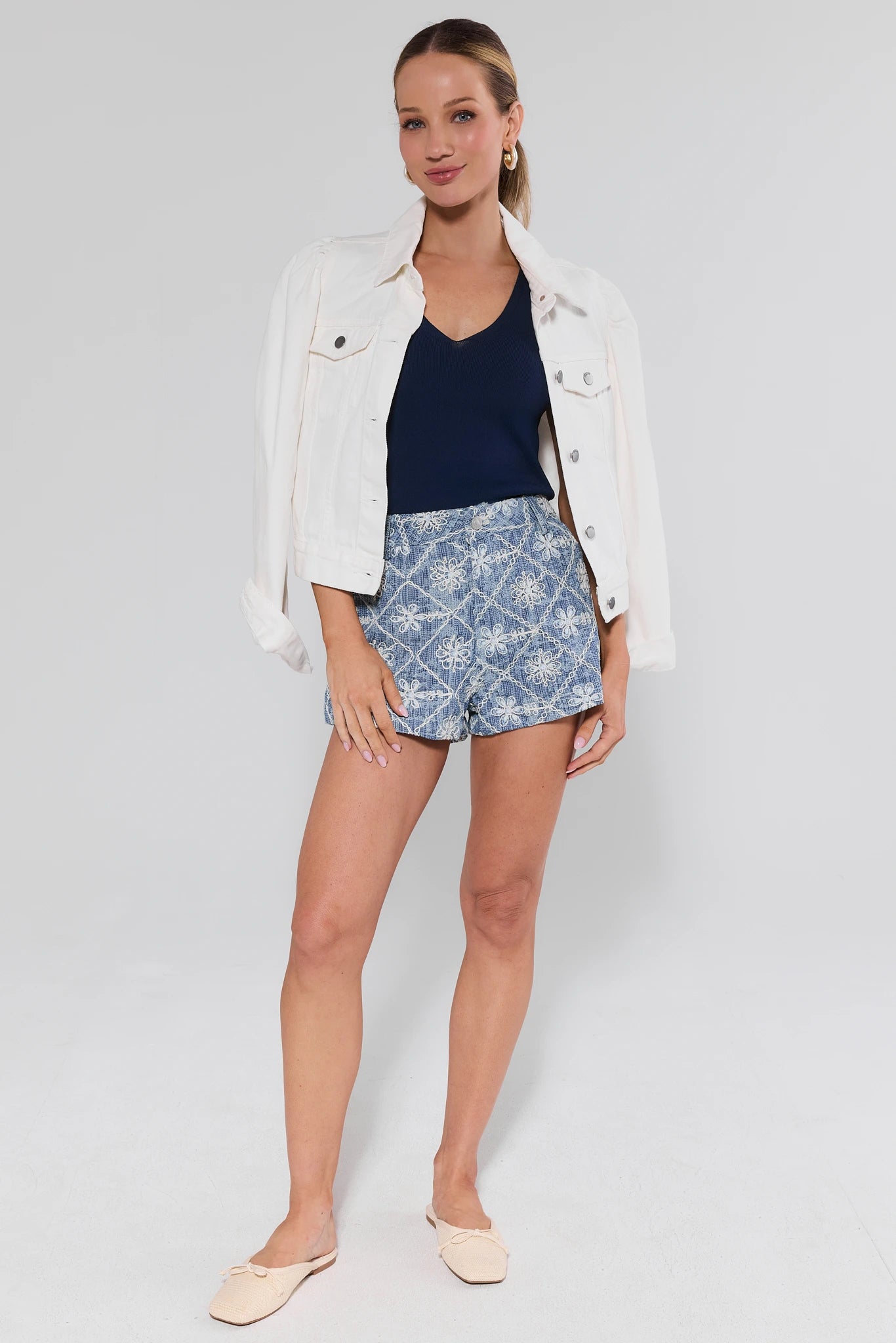 Woman wearing a white jacket, navy top, and floral shorts on a plain background