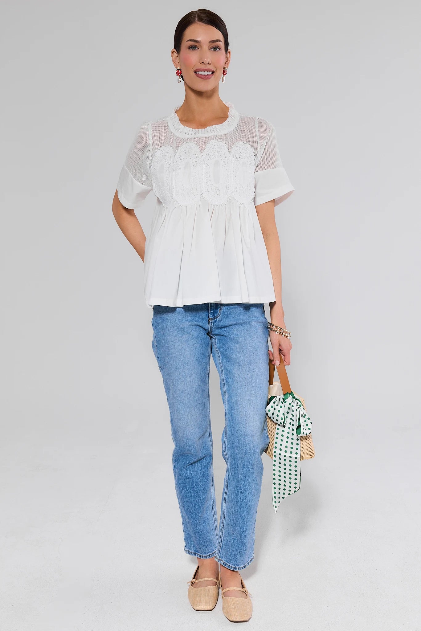 Woman wearing a white blouse with lace details and blue jeans, standing against a plain background.