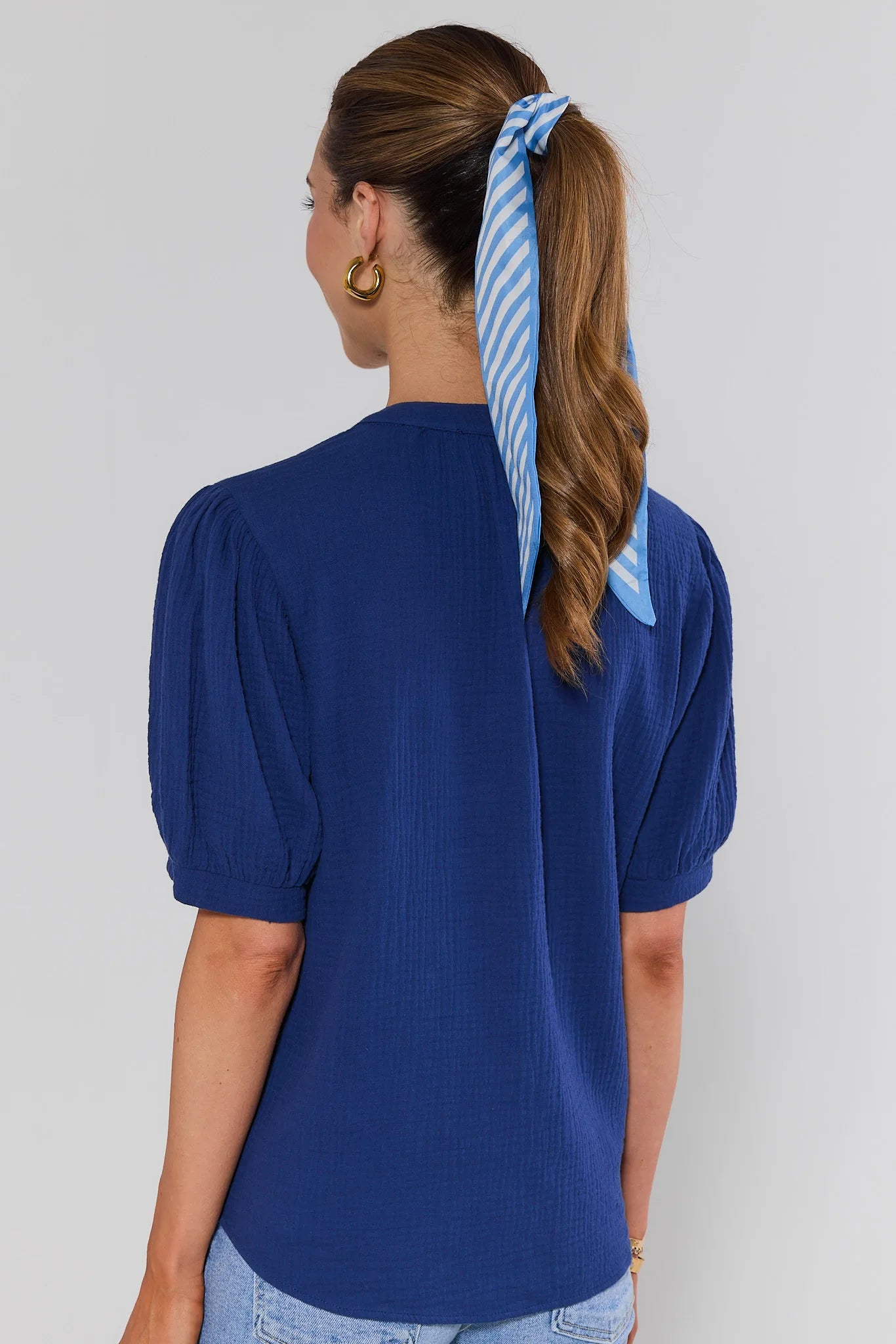 Woman standing wearing a blue top with puffed sleeves and a blue and white striped hair tie in her hair against a plain background.