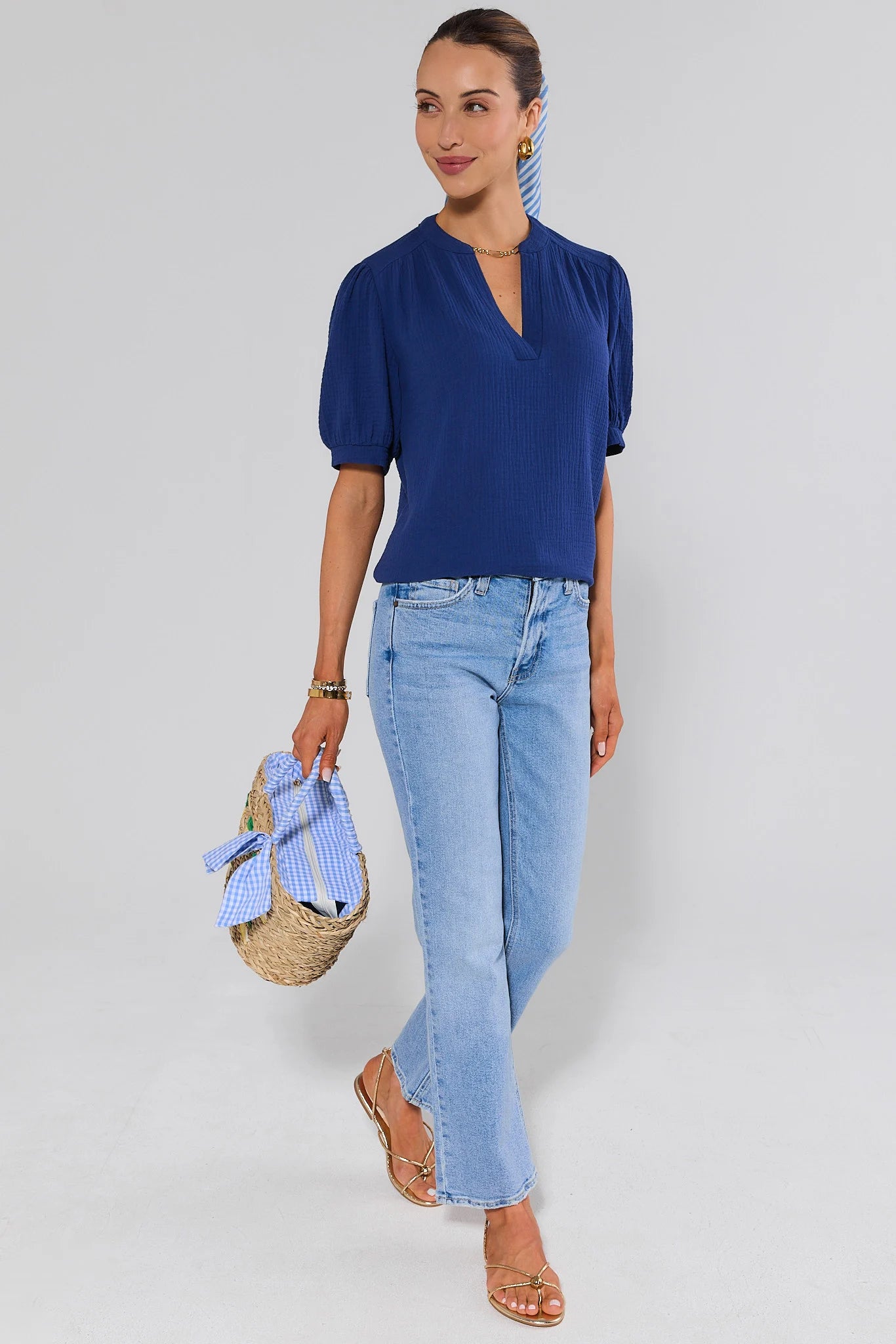 Woman wearing a blue top and light blue jeans, holding a straw bag against a white background