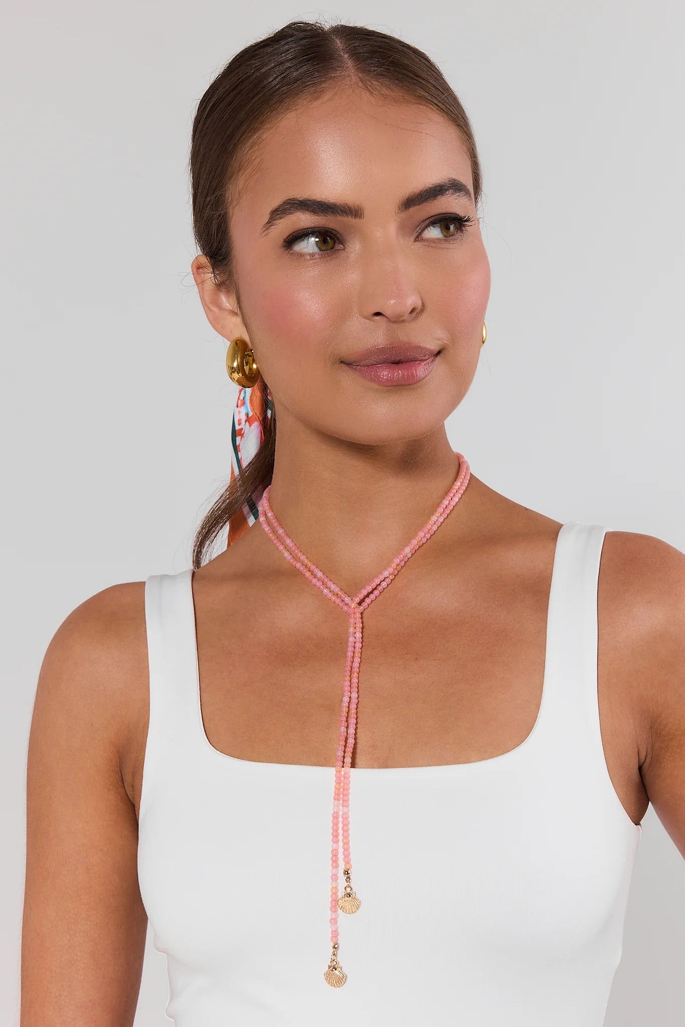 Woman wearing a pink beaded necklace with a white top on a plain background