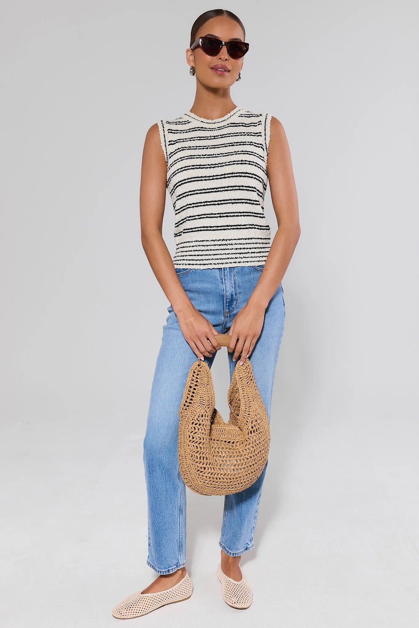 Woman wearing a striped sleeveless top and blue jeans, holding a woven bag on a white background