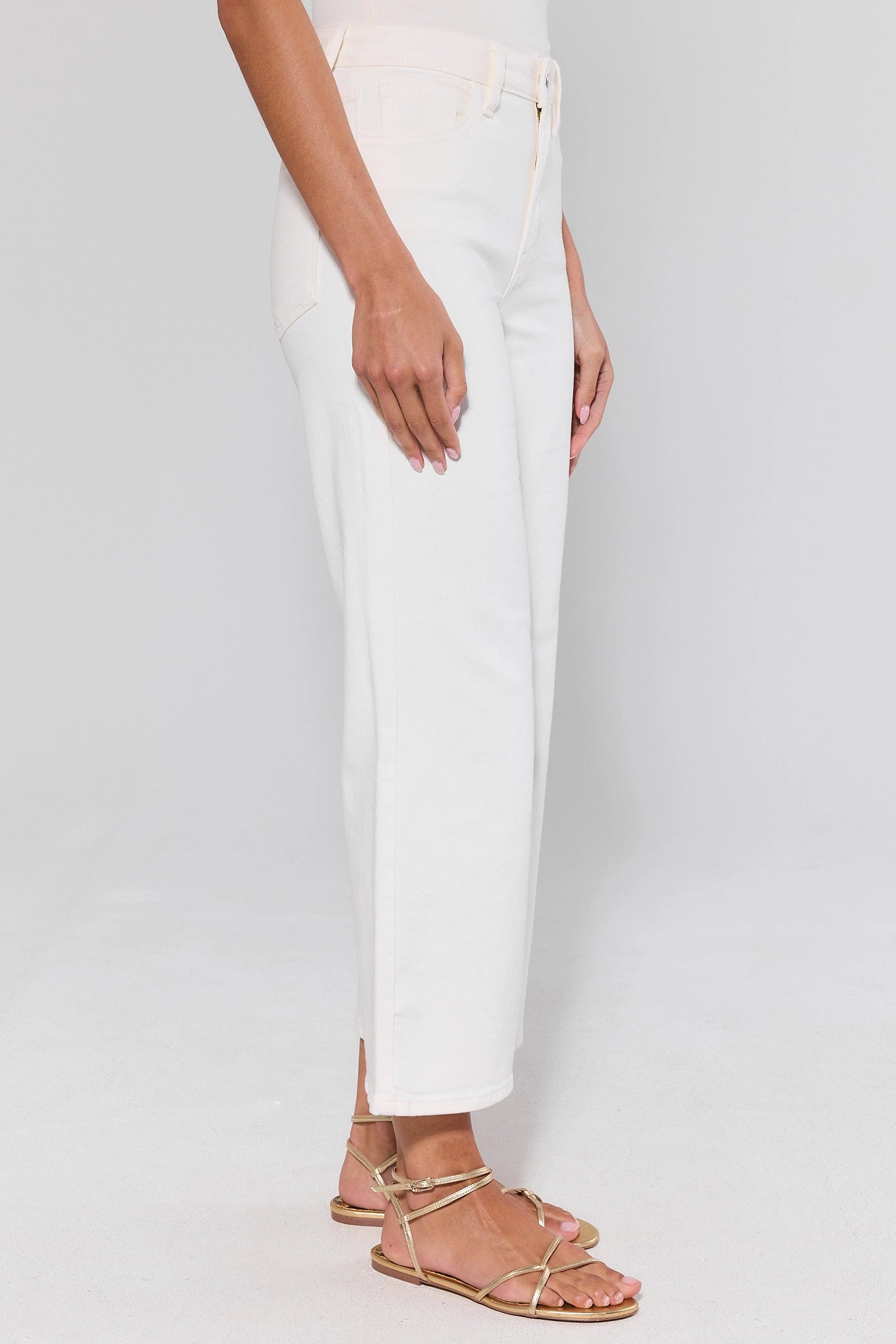 side shot Person wearing white pants and gold sandals on a plain background