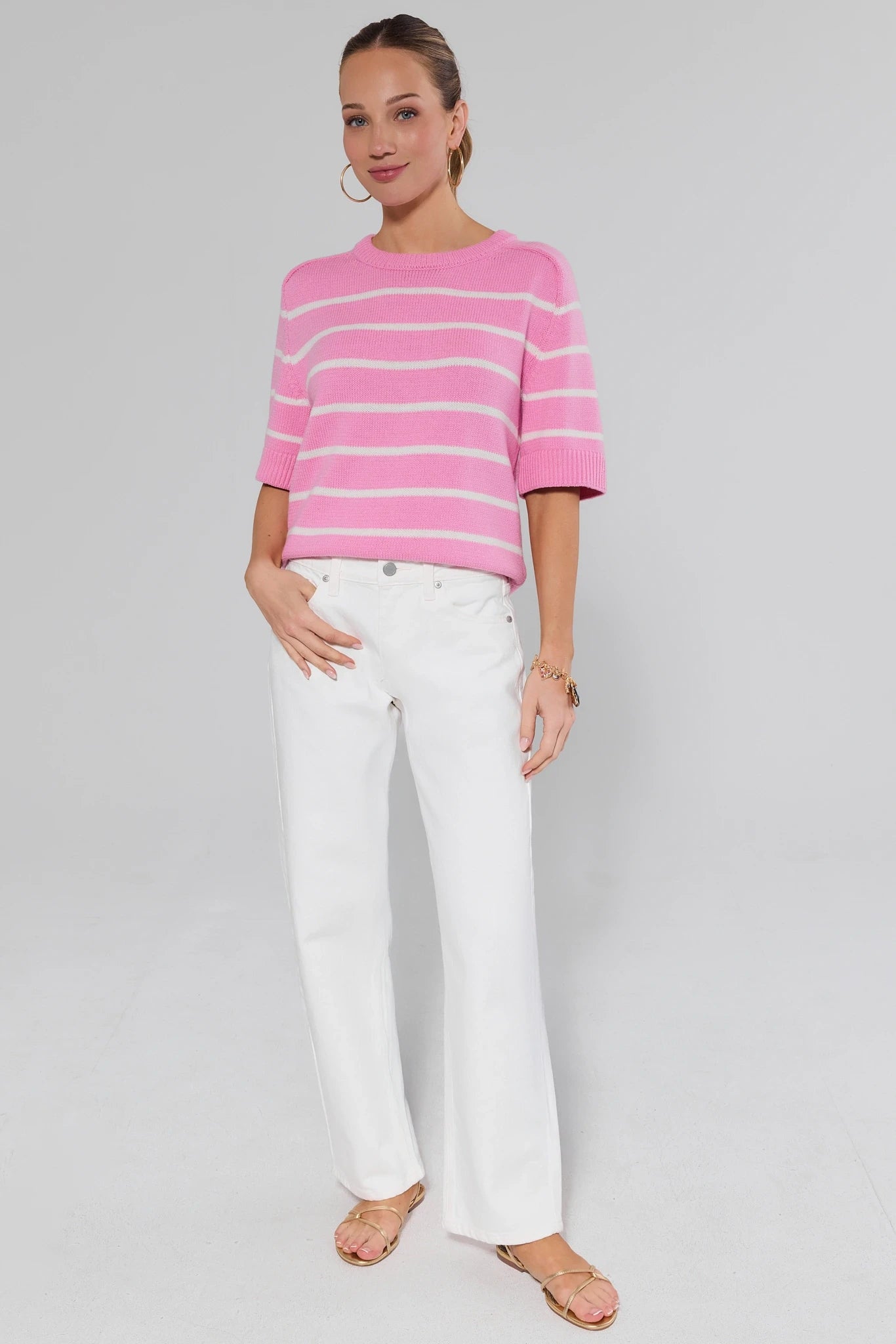 Woman wearing a pink and white striped sweater and white pants on a gray background