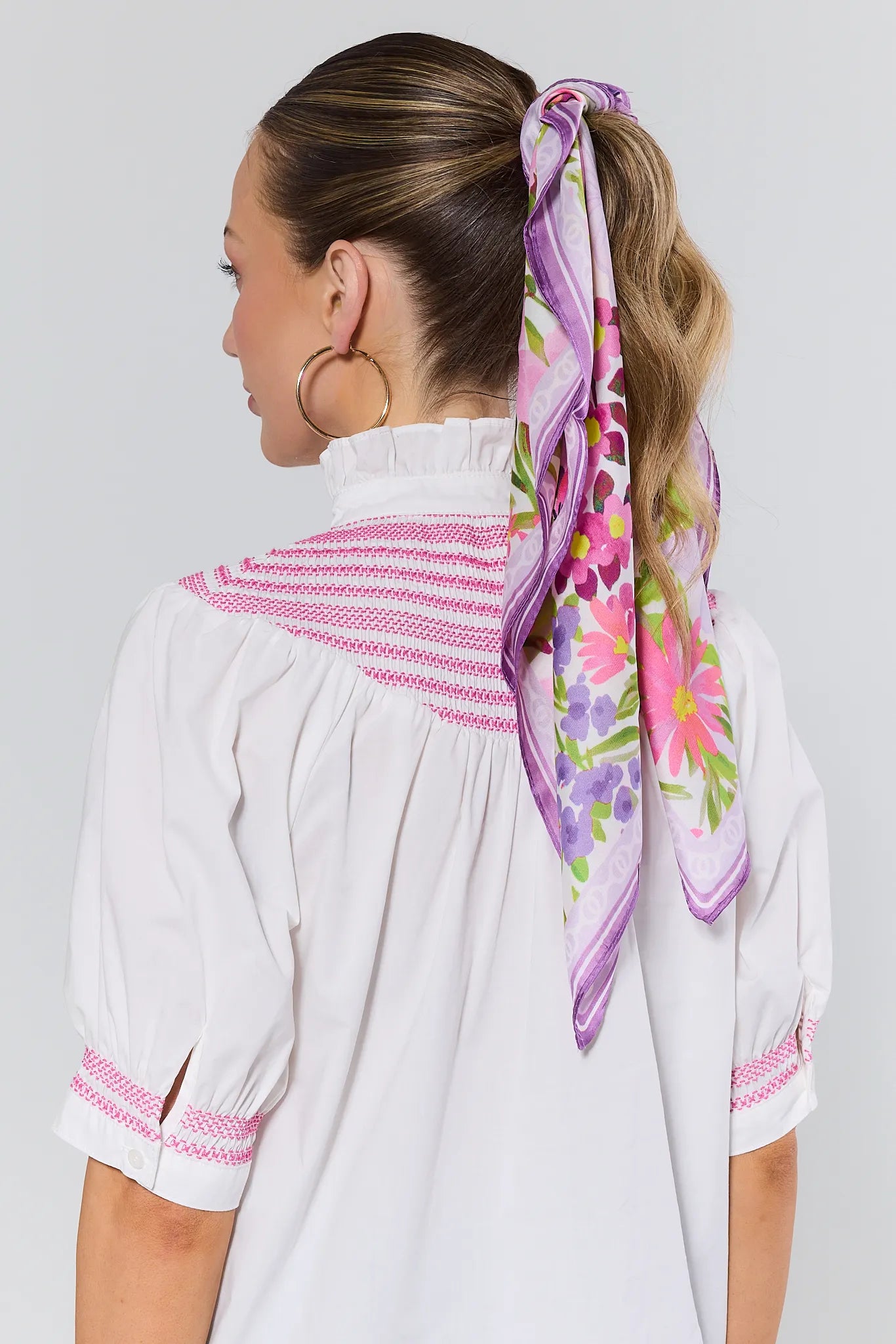 Woman wearing a white blouse with pink trim and a colorful floral scarf in her hair against a plain background