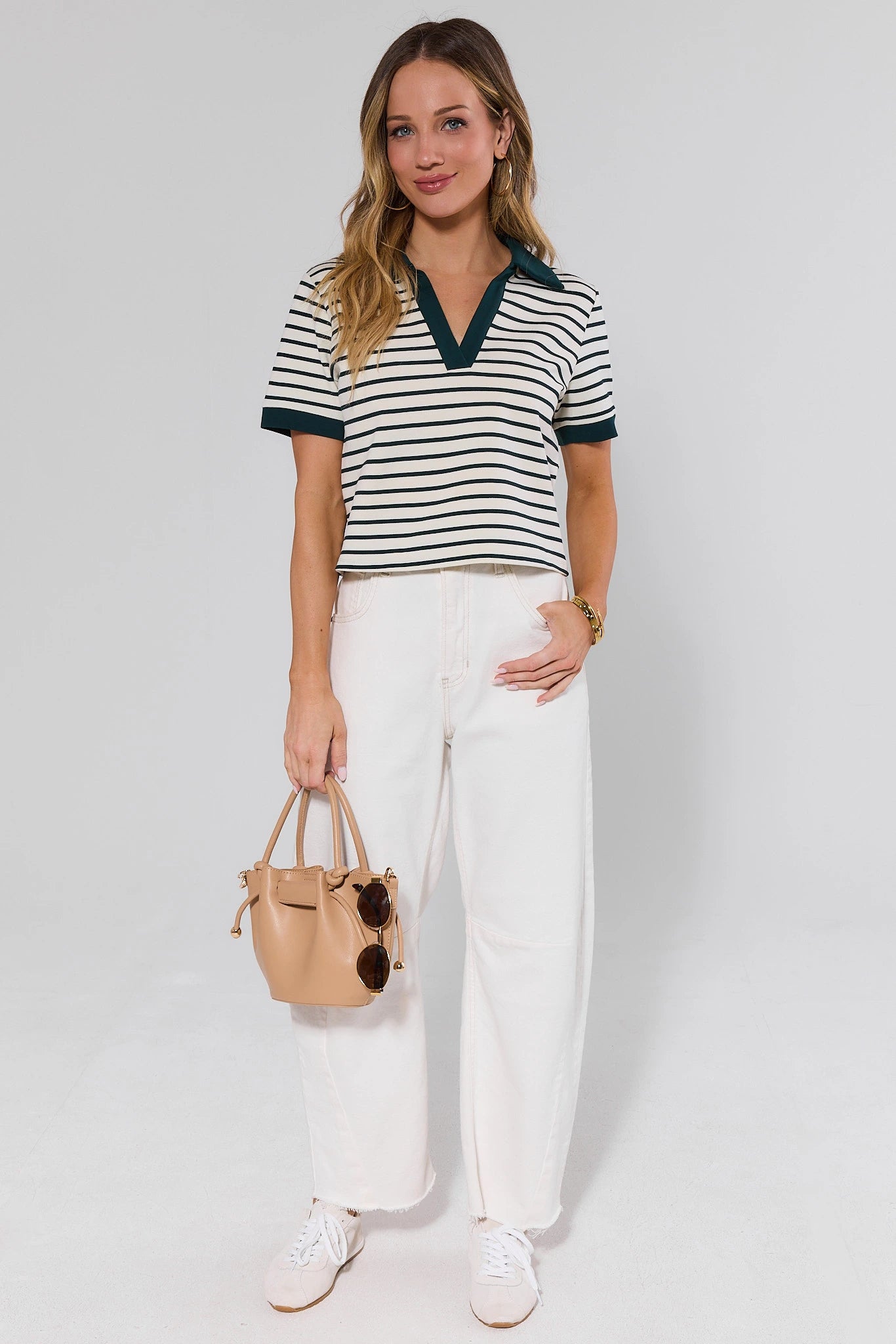 Woman wearing a striped shirt and white pants with a beige handbag on a plain background