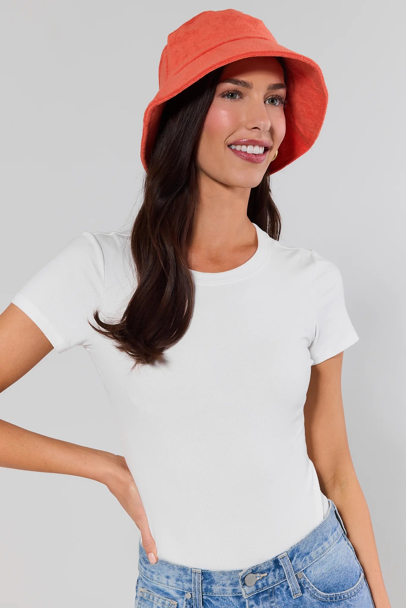 Woman wearing a orange bucket hat, white t-shirt, and blue jeans on a gray background