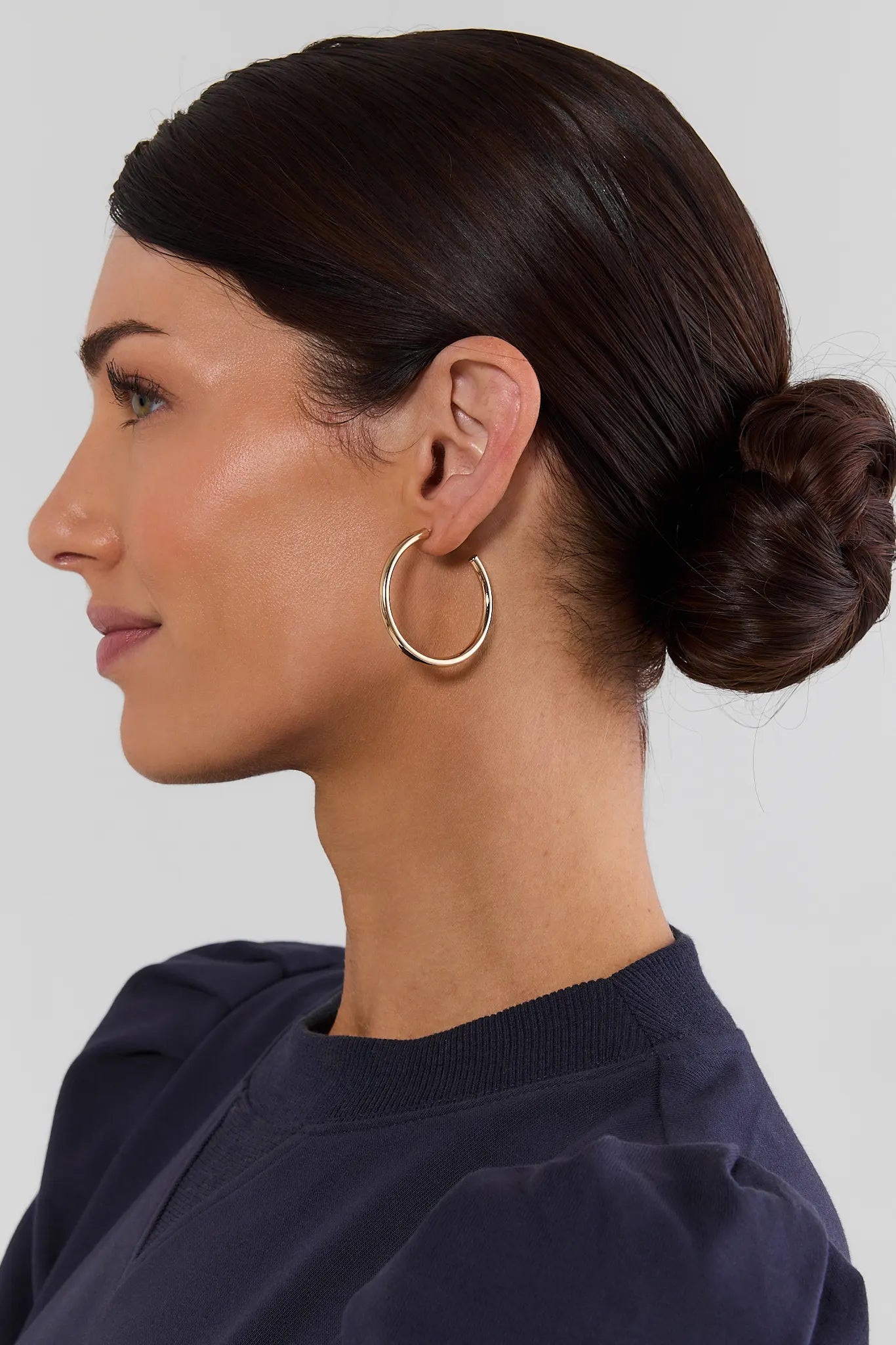 A woman in profile wearing a pair of large, thin gold hoop earrings, with her dark hair styled in a low bun and wearing a navy blue top against a light gray background.