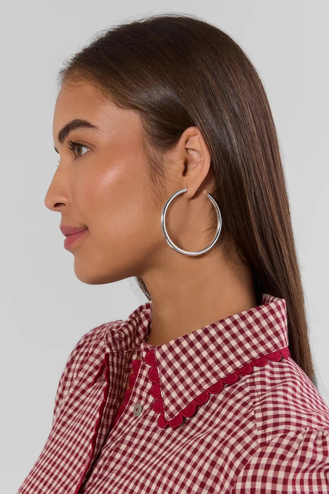 A woman shown in profile wearing a large silver-toned hoop earring and a red-and-white gingham shirt with red trim, against a light gray studio background.