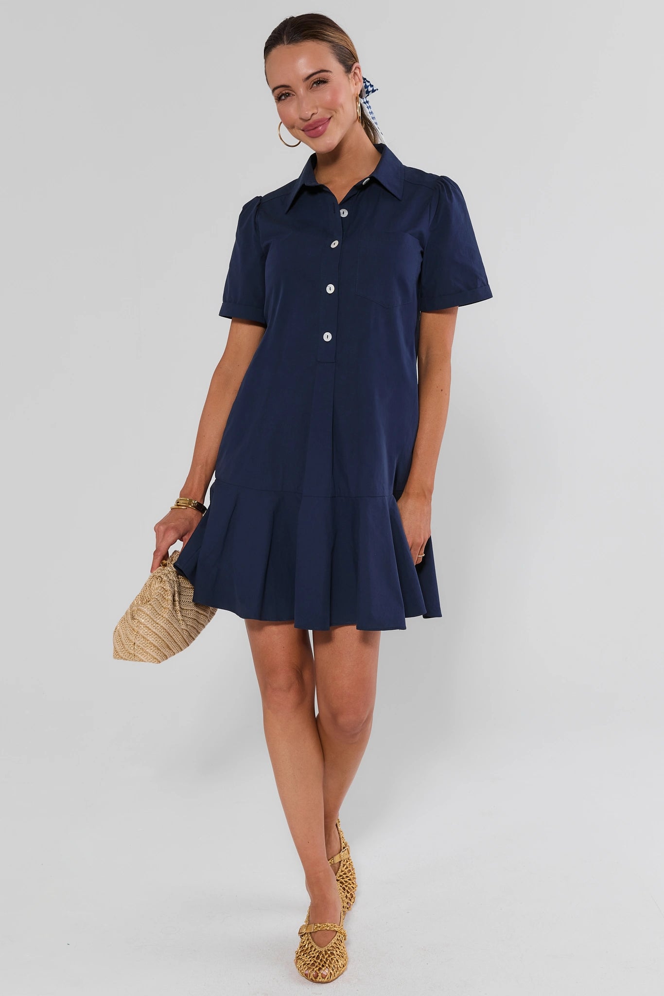 A woman modeling a short-sleeve navy blue collared shirt dress with a button-front bodice and flared skirt, standing against a light gray studio background and holding a woven straw handbag.