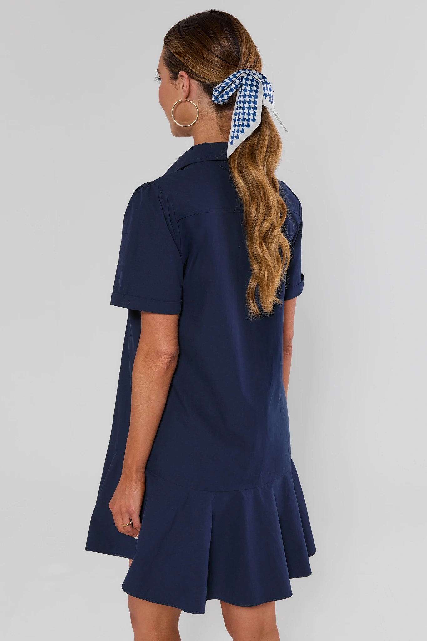 A woman shown from the back wearing a navy blue short-sleeve collared dress with a flared hem, paired with a blue and white gingham hair tie.