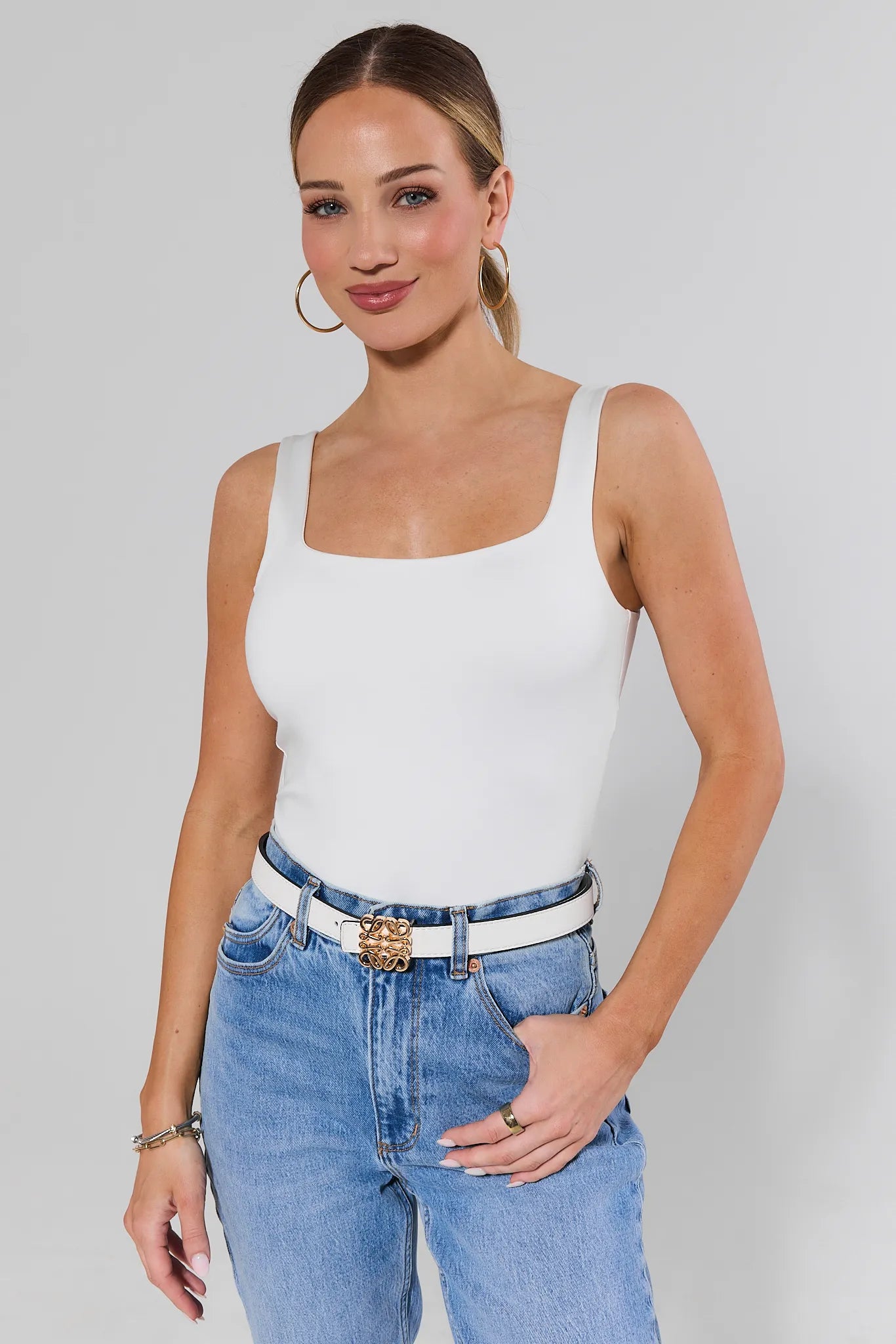 Woman wearing a white tank top and blue jeans white rowan belt against a plain background