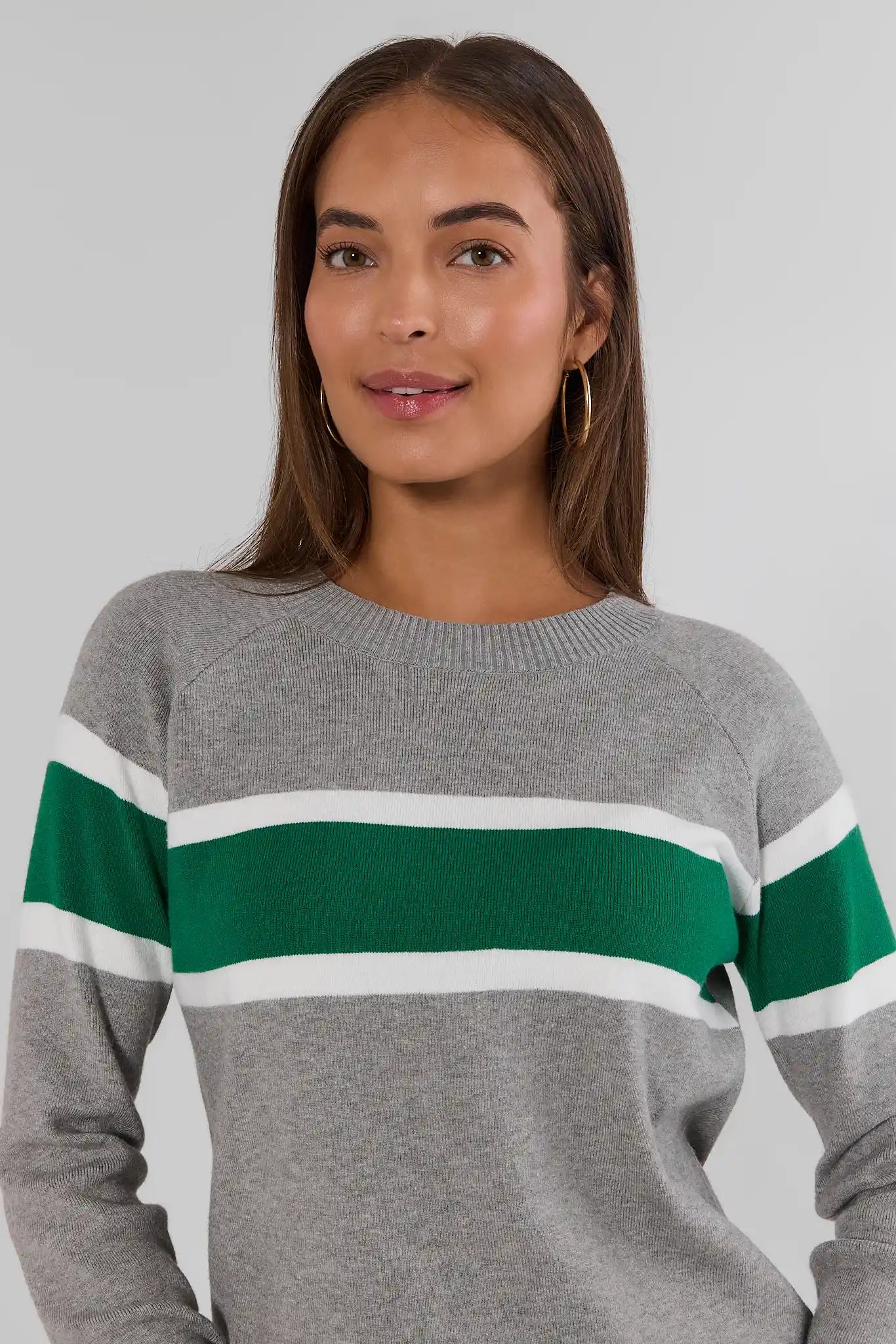 A woman wearing a gray crewneck sweater with a wide green and white horizontal stripe across the chest, photographed against a light gray studio background.