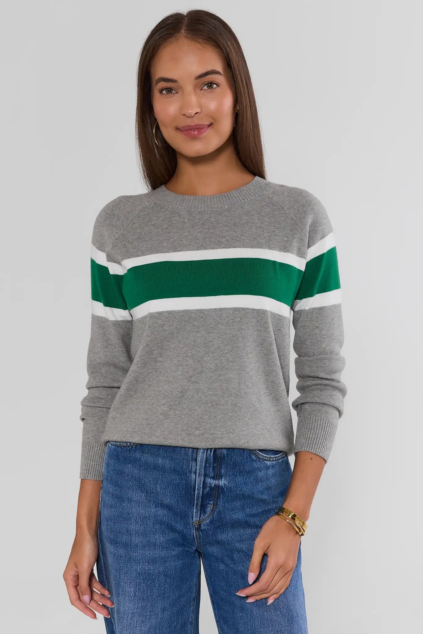 A woman wearing a gray knit sweater with horizontal green and white stripes across the chest, paired with blue jeans, photographed against a light gray studio background.