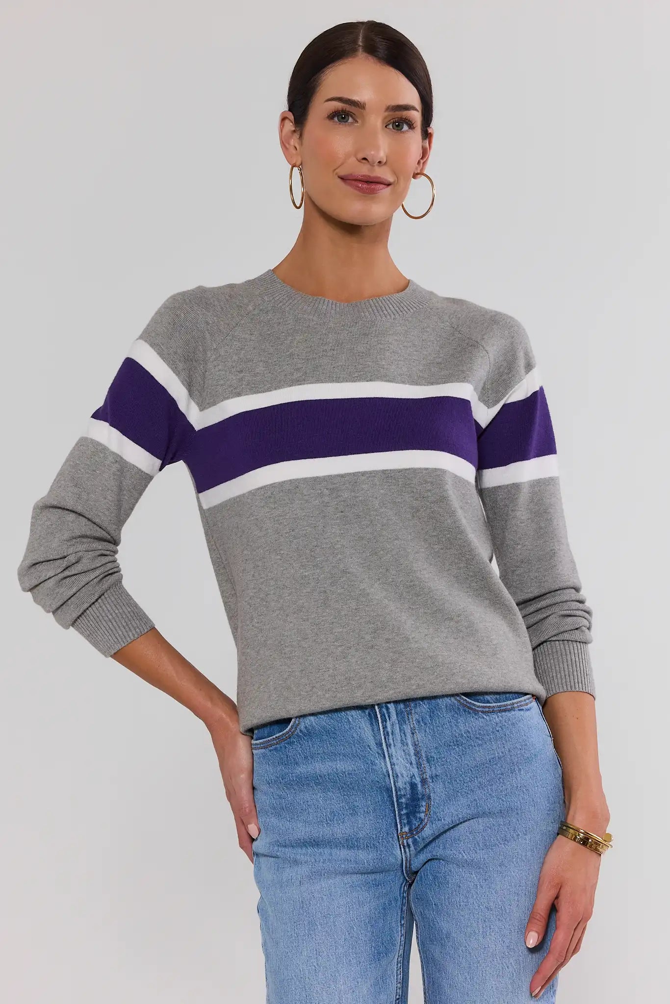A woman wearing a gray knit sweater with horizontal purple and white stripes across the chest and sleeves, paired with light blue jeans, against a light gray studio background.