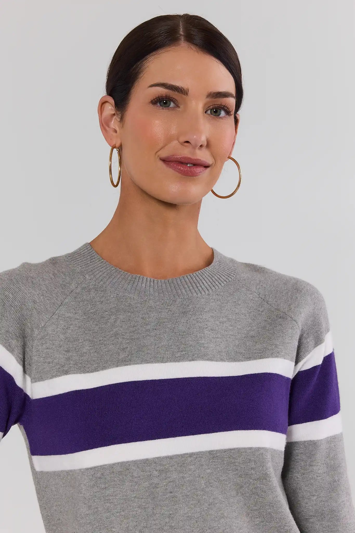 A woman wearing a gray knit sweater with horizontal purple and white stripes, paired with gold hoop earrings, against a light gray background.