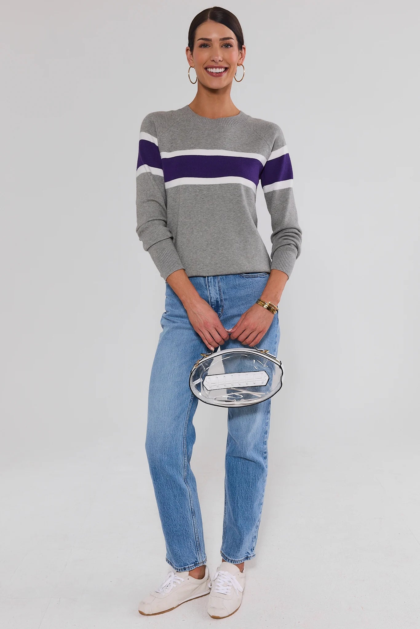 A woman modeling a gray crewneck sweater with purple and white horizontal stripes on the chest and sleeves, paired with light blue jeans, white sneakers, and holding a clear clutch bag.