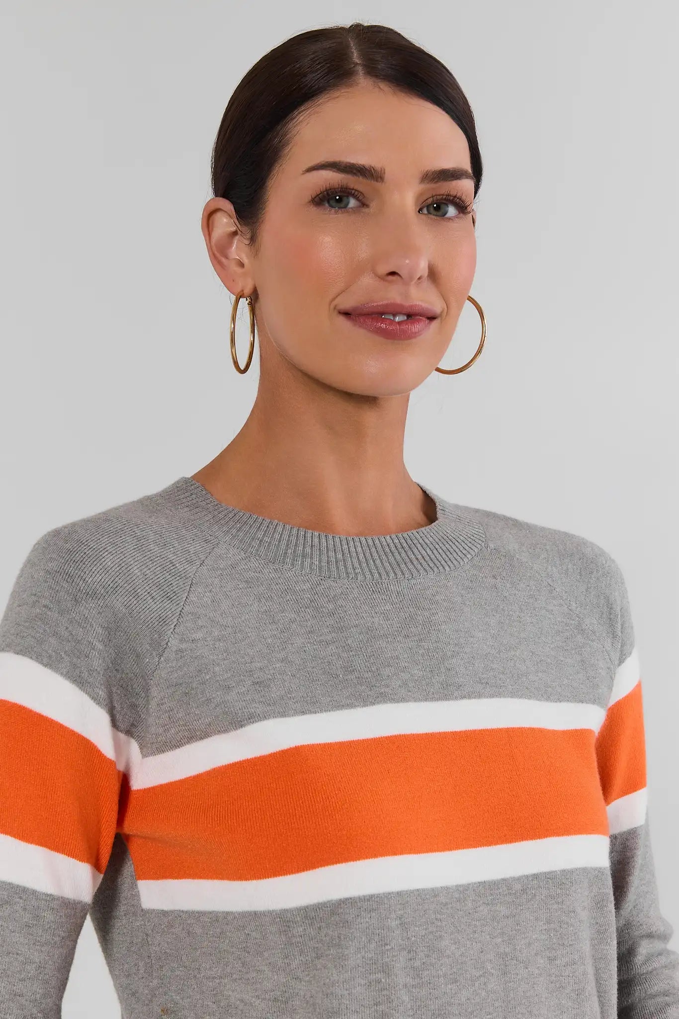 A woman wearing a heather gray knit sweater with bold horizontal orange and white stripes across the chest, paired with large gold hoop earrings, against a plain light gray studio background.