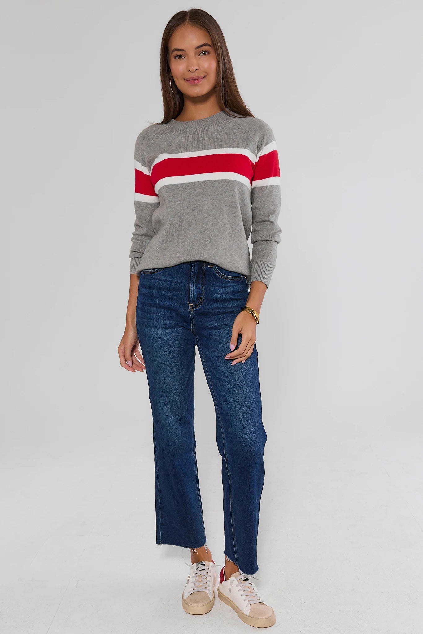 A woman modeling a gray knit sweater with bold red and white horizontal stripes across the chest and shoulders, paired with dark blue cropped jeans and beige sneakers, against a light gray studio background.