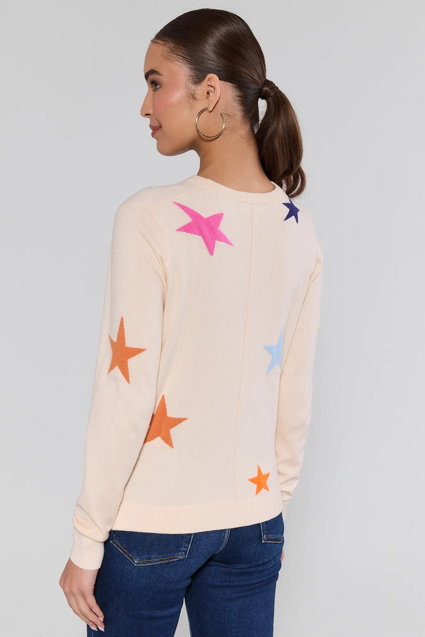Woman wearing a cream sweater with colorful star patterns and blue jeans on a plain background Luna Cream Star Knit Sweater