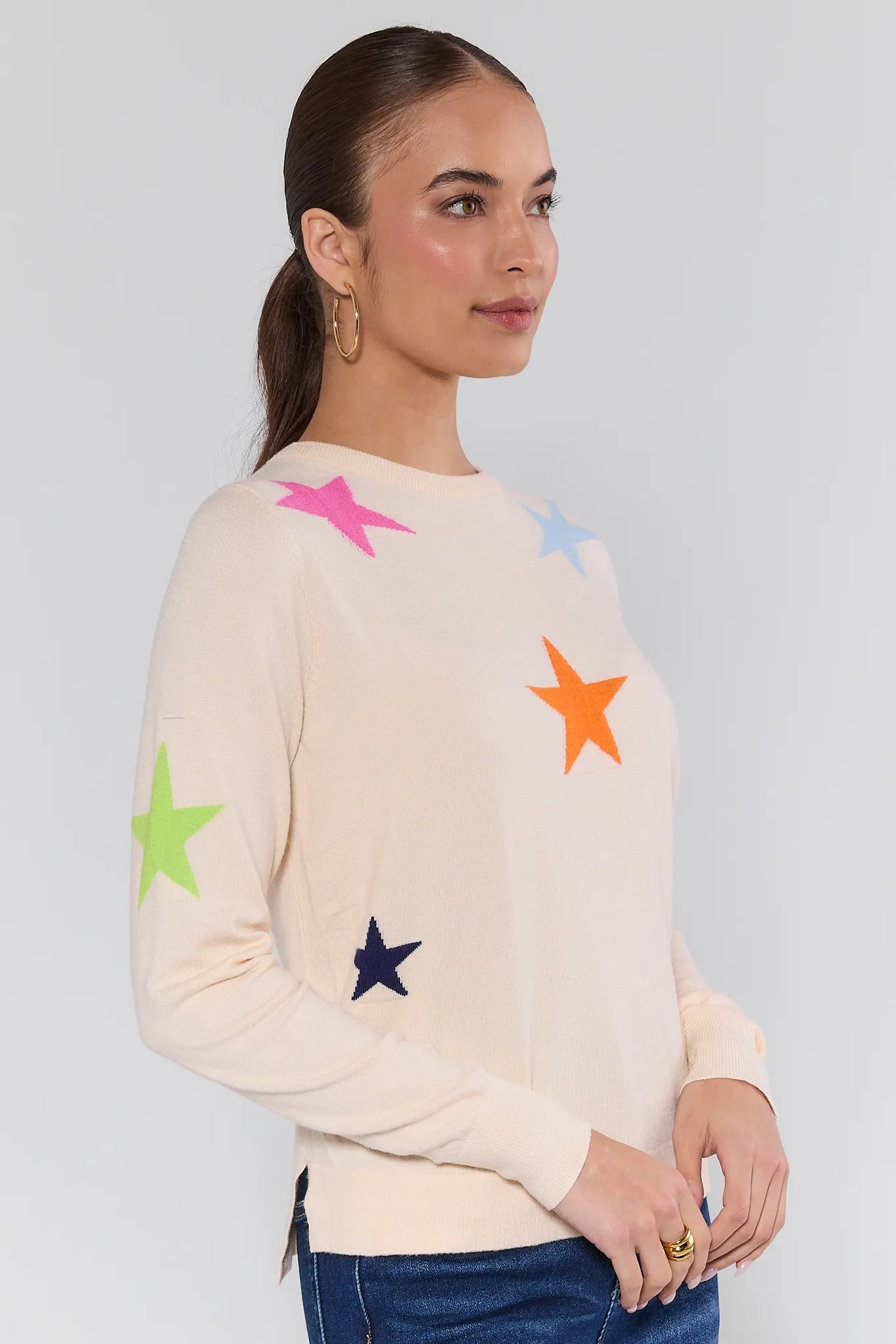 Woman wearing a cream sweater with colorful star patterns and blue jeans on a plain background Luna Cream Star Knit Sweater