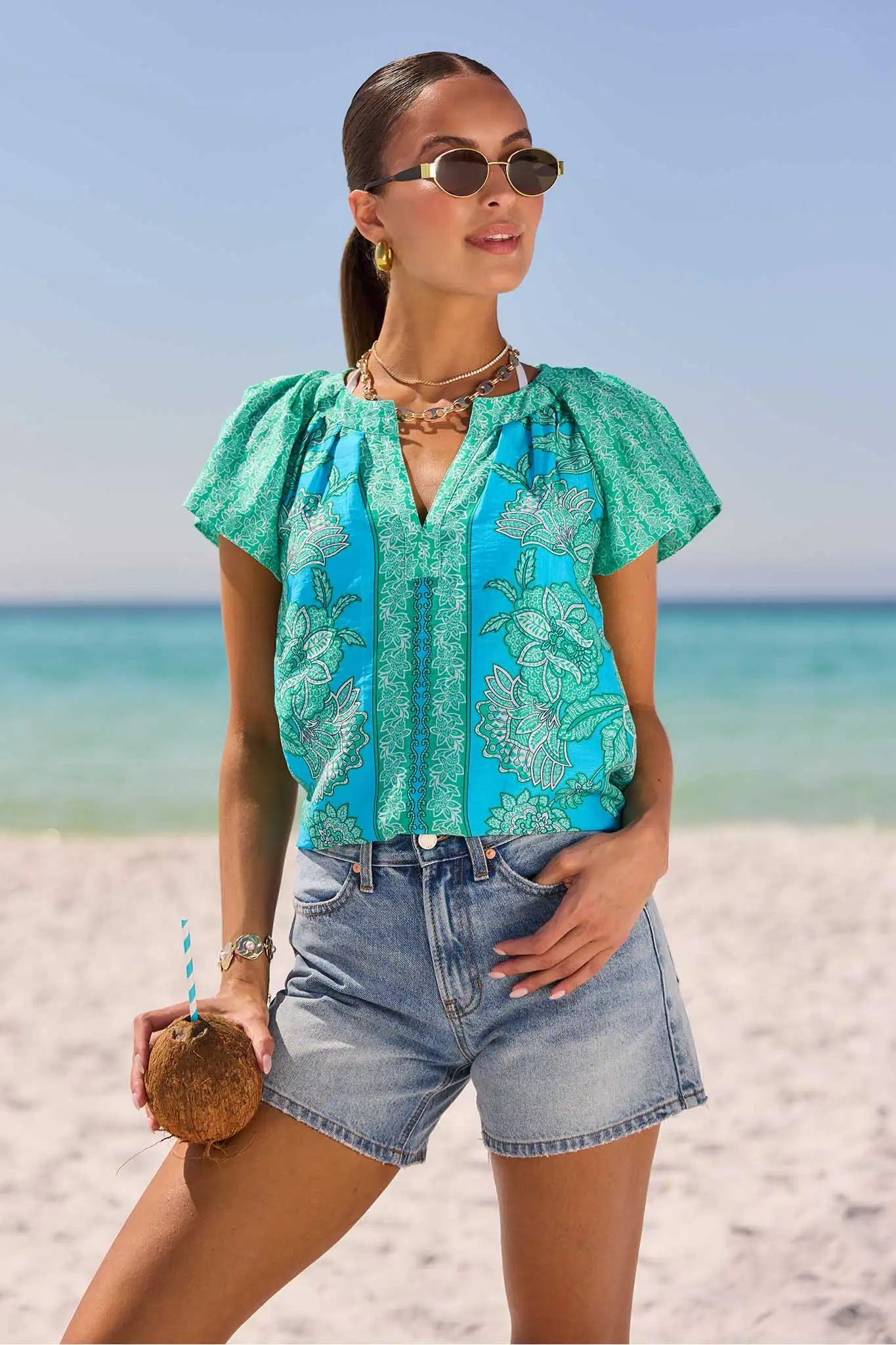 Woman in a turquoise top and denim shorts standing on a beach with ocean view.