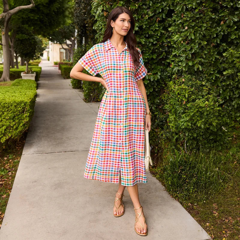 Woman in a colorful checkered dress standing on a sidewalk with greenery around. collection