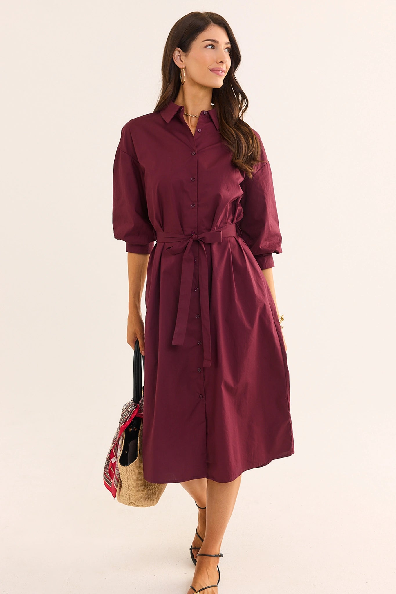 A woman modeling a burgundy midi shirt dress with three-quarter sleeves, a button-down front, and a matching waist tie, holding a patterned tote bag against a plain off-white background.