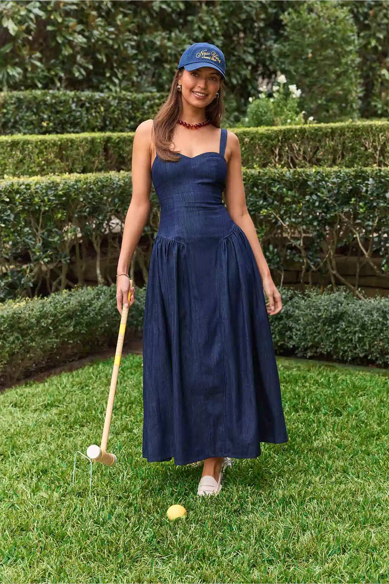 Woman in a navy dress and cap playing croquet on grass