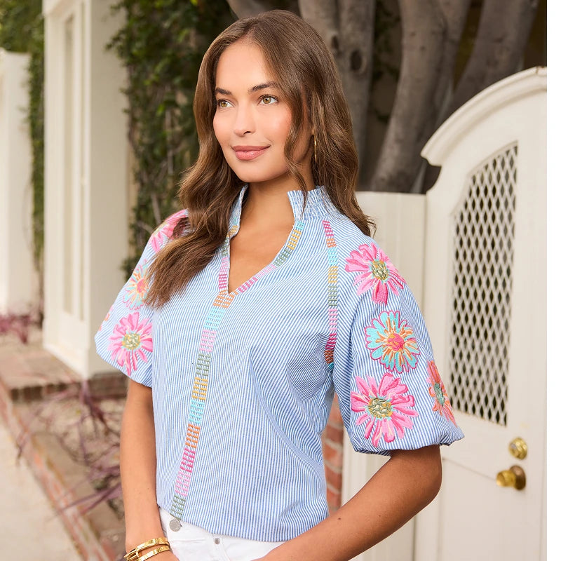 Woman wearing a blue blouse with colorful floral embroidery in an outdoor setting. collection