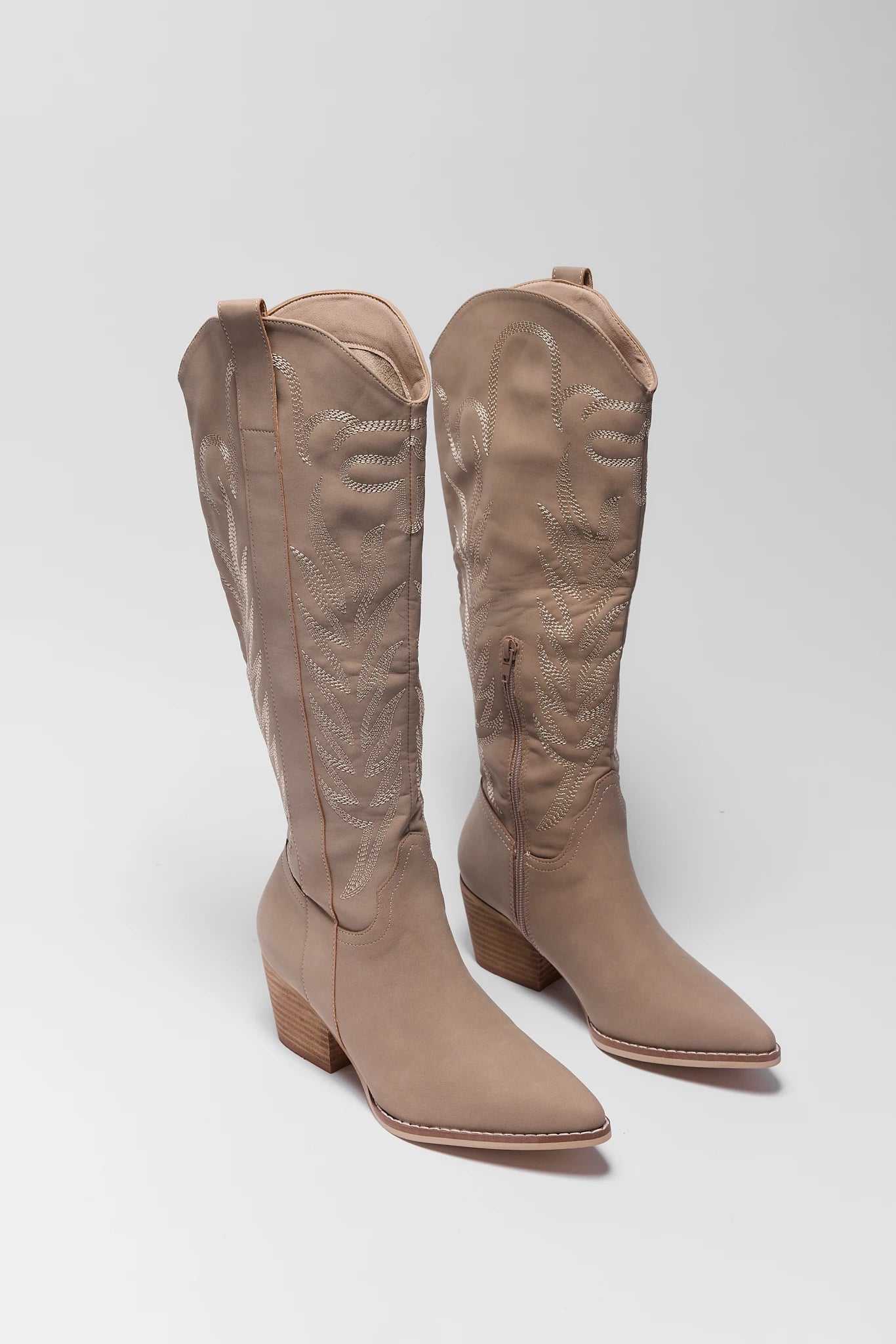 A pair of knee-high western boots in a light taupe-beige color, featuring embossed floral detailing on the shafts and low block heels, shown against a neutral light gray background.